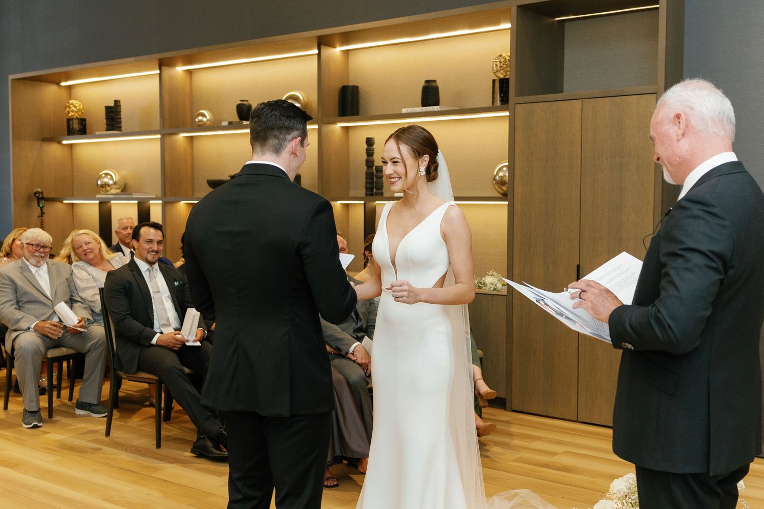Wedding guests dance and socialize in an elegant wood-paneled reception venue with warm lighting.