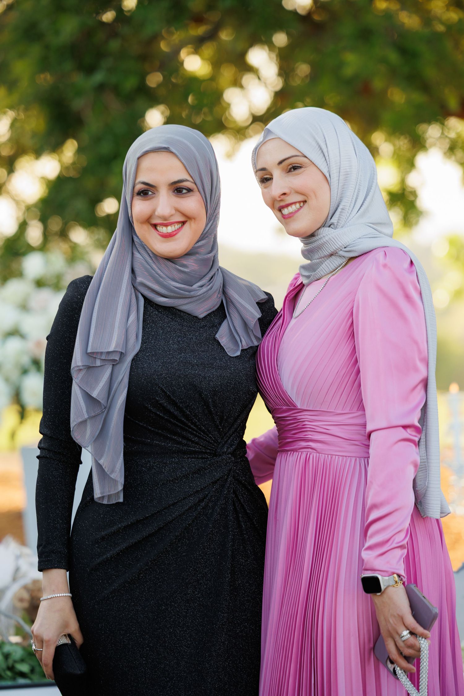 Two people wearing black and pink dresses with grey hijabs share a candid moment outdoors in natural lighting.