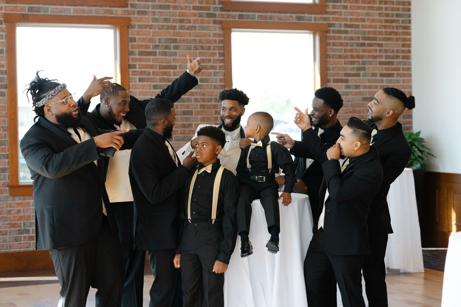 A group wearing formal black suits and ties gather together in celebration at an indoor venue with brick walls and windows.