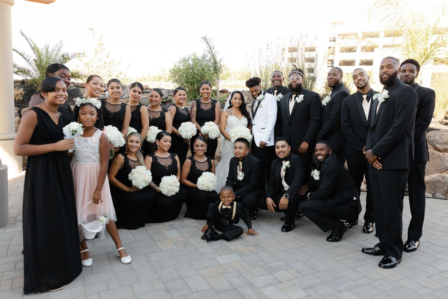 Large wedding party in formal black and white attire poses together on outdoor steps for group photo.