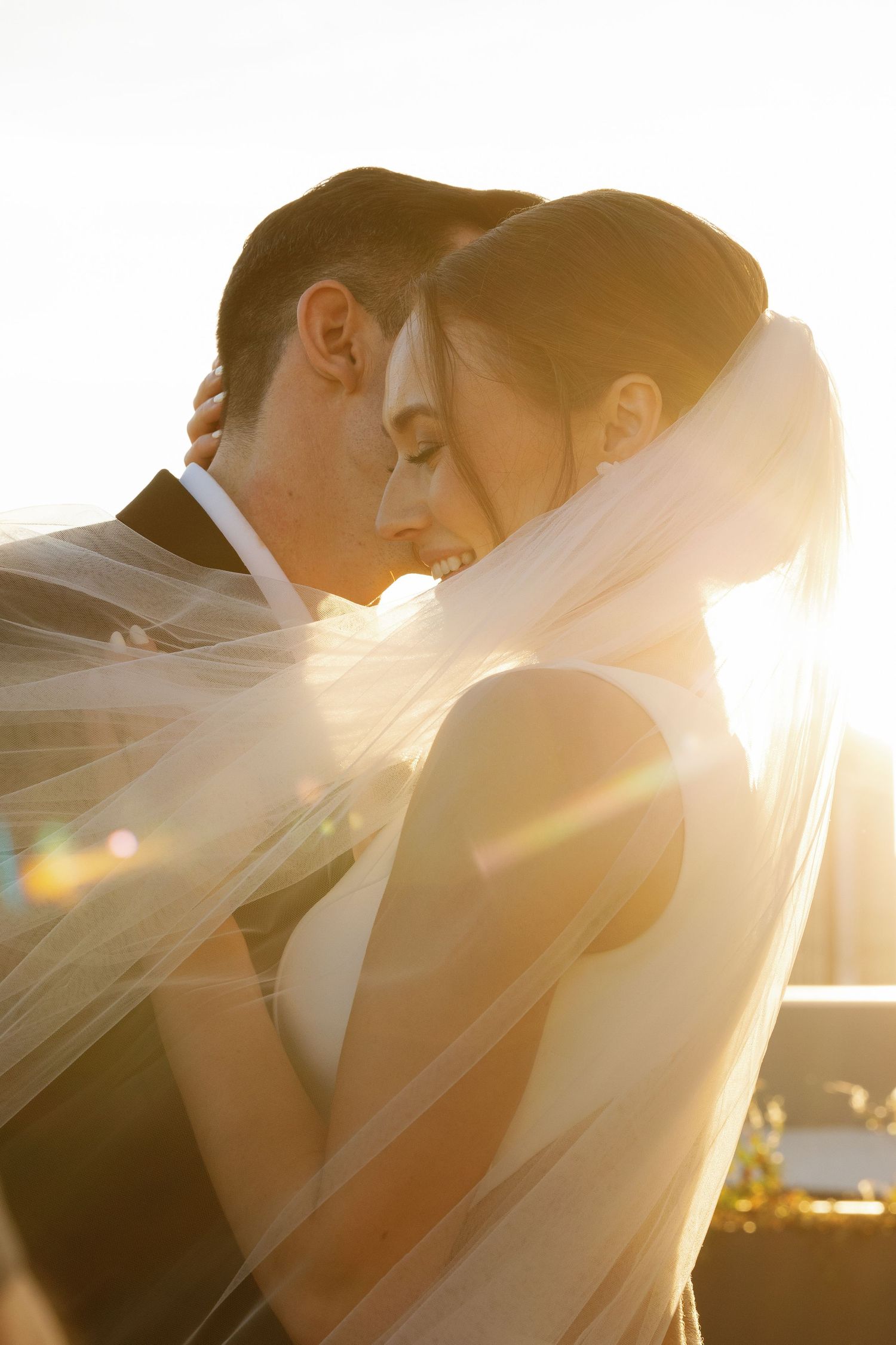 A romantic sunset silhouette of a couple embracing under a flowing bridal veil with golden sunlight streaming through.