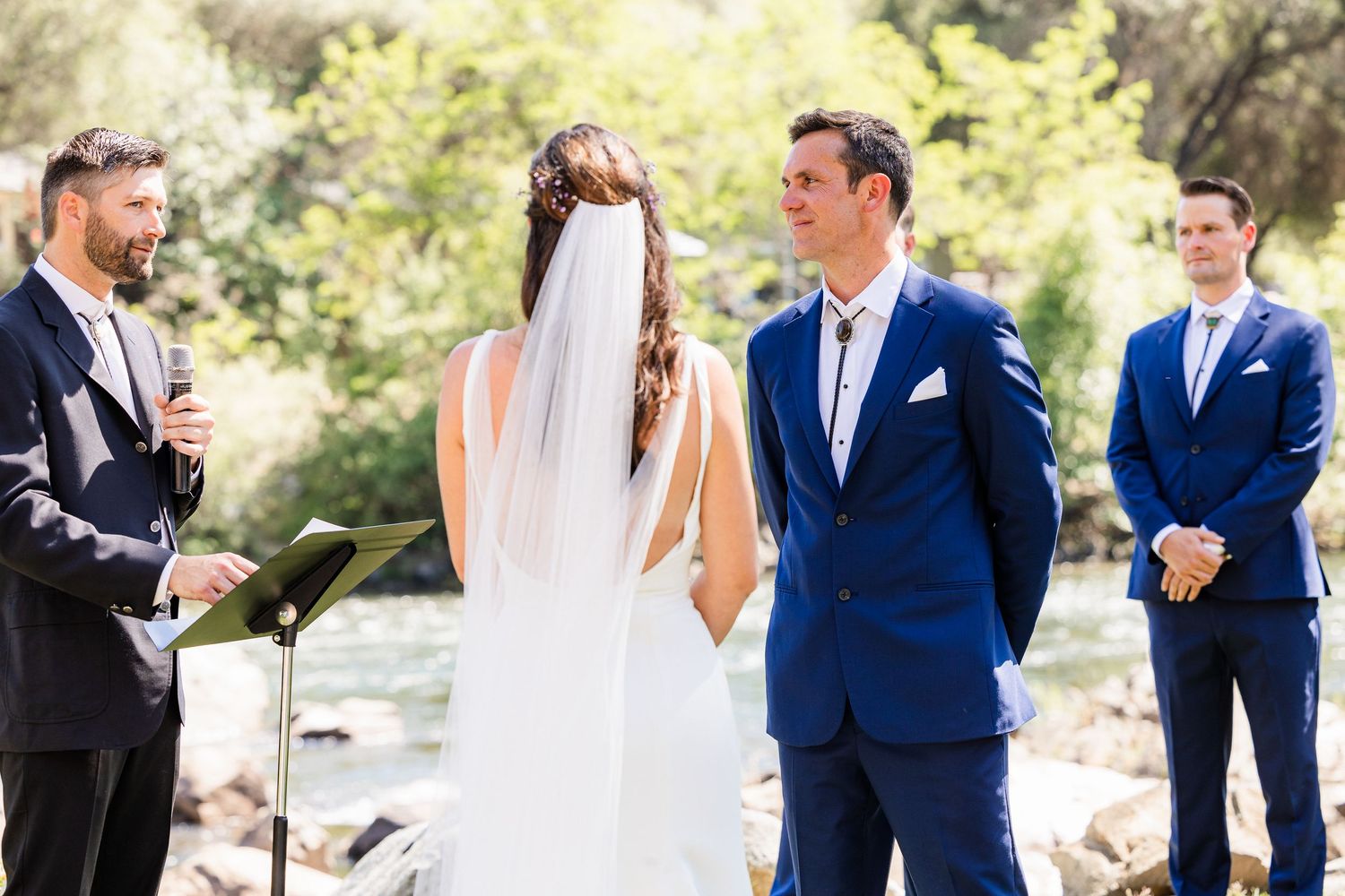 Wedding party in navy blue suits and white dresses celebrates outdoors during a sunny garden ceremony.