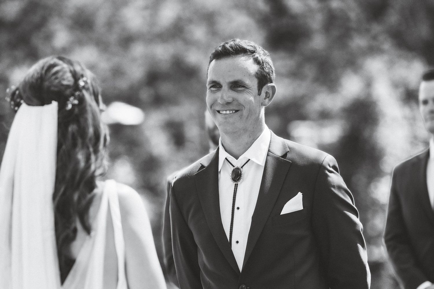 Black and white photo of a wedding party standing in a row outdoors with guests in formal attire.