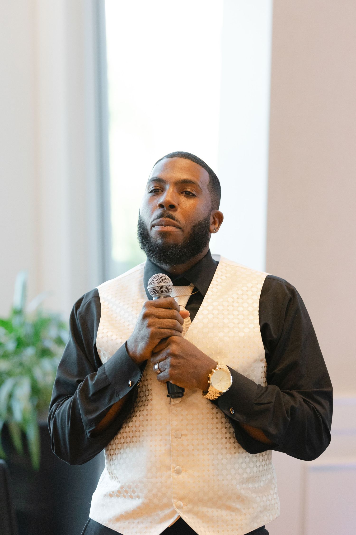A speaker in a white vest and black shirt holds a microphone while presenting at an indoor event.