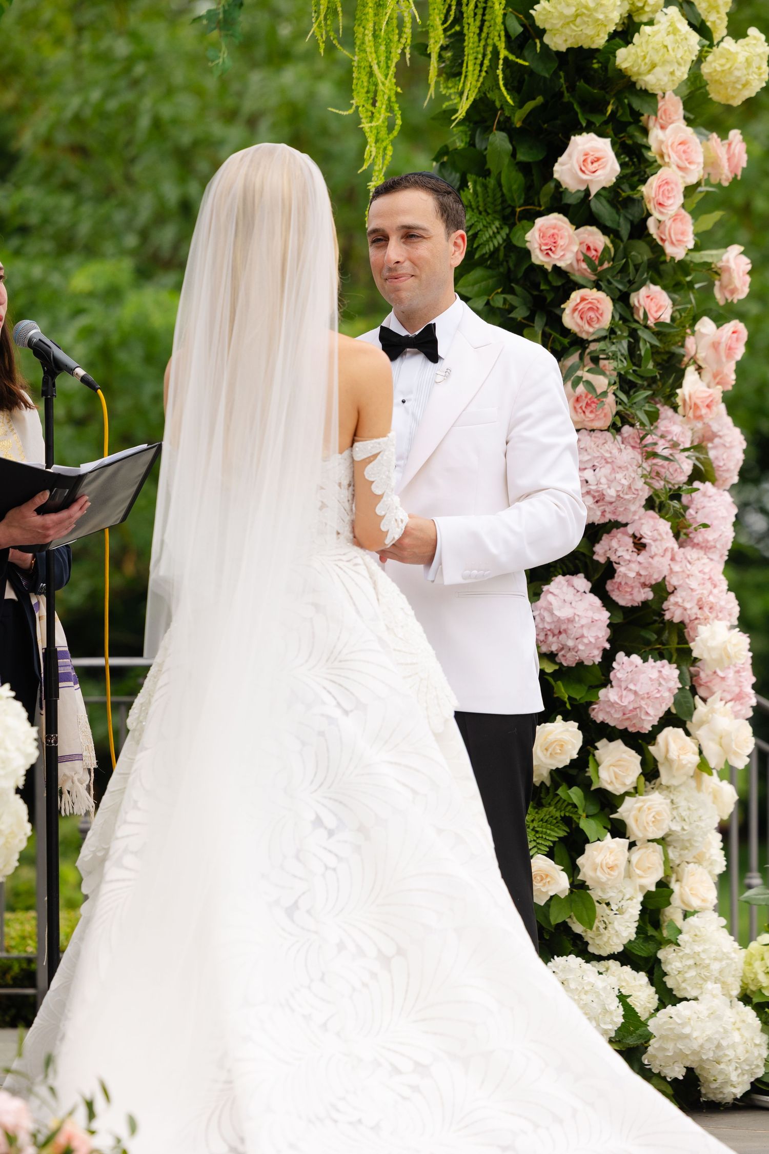 A wedding couple exchanges vows under an elegant floral arch adorned with pink and white roses and hydrangeas.