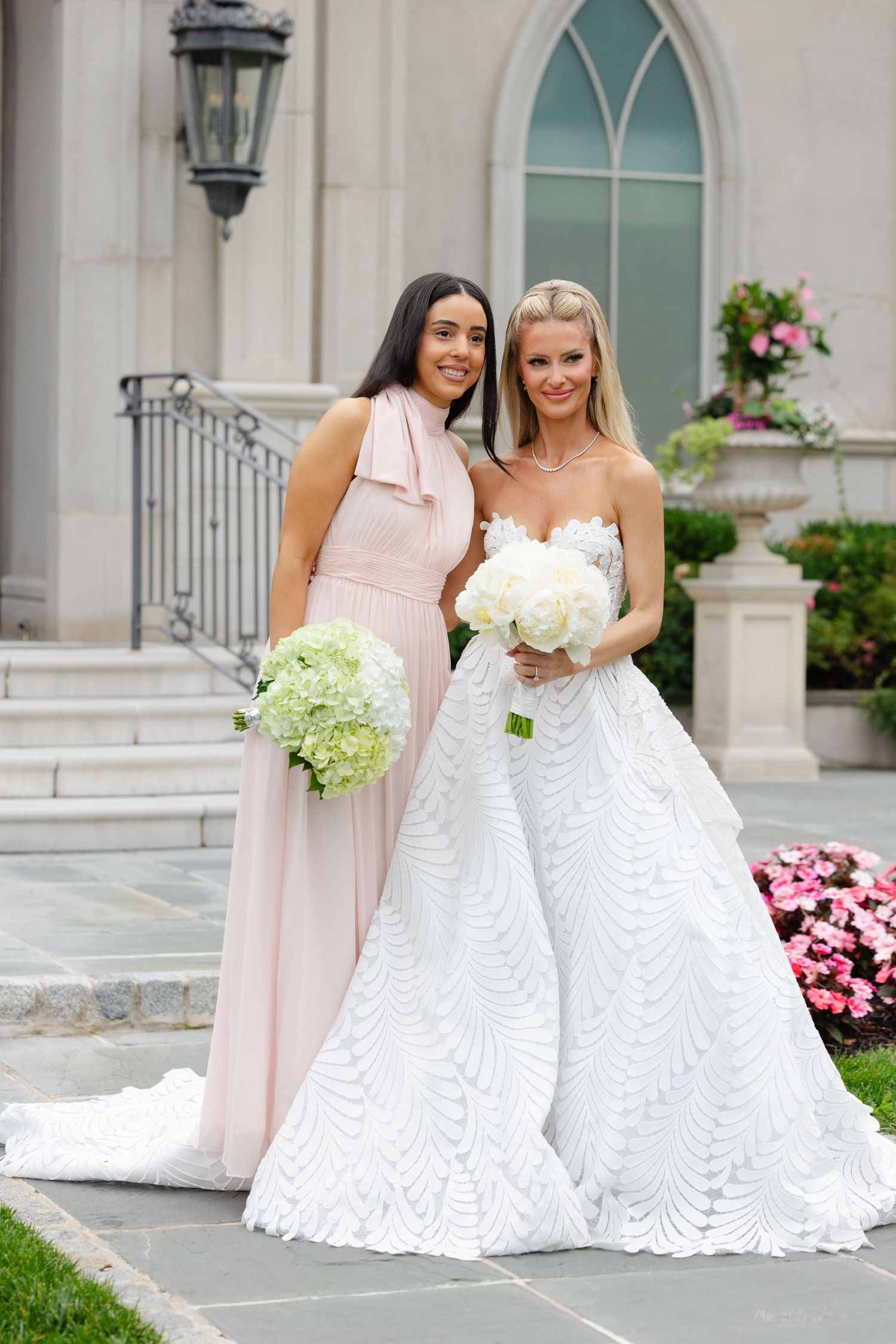 A bride in a white lace gown and bridesmaid in blush pink dress pose together outside a church with floral decor.