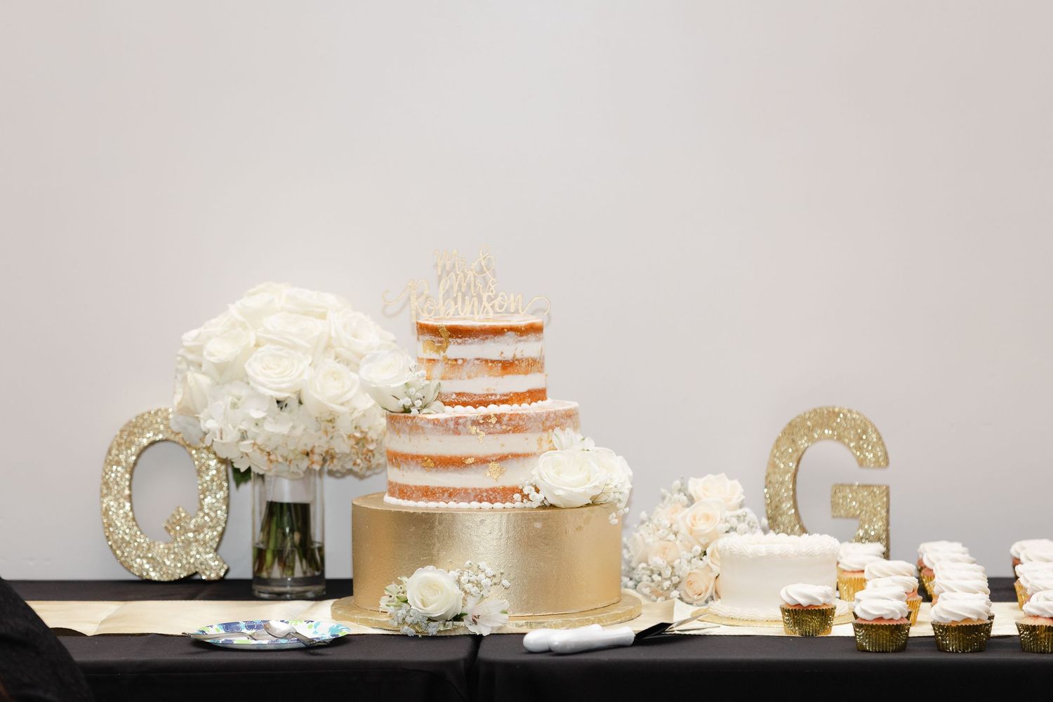 An elegant wedding dessert table features a naked cake, white floral arrangements and gold letter decorations.