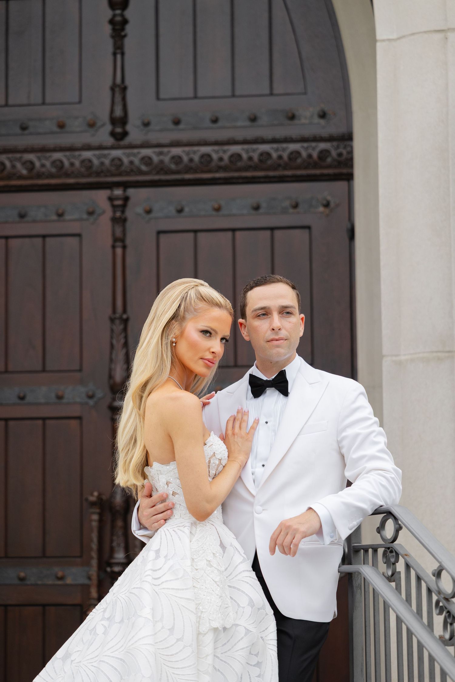 A couple in formal wedding attire poses on white stairs in front of ornate wooden doors.