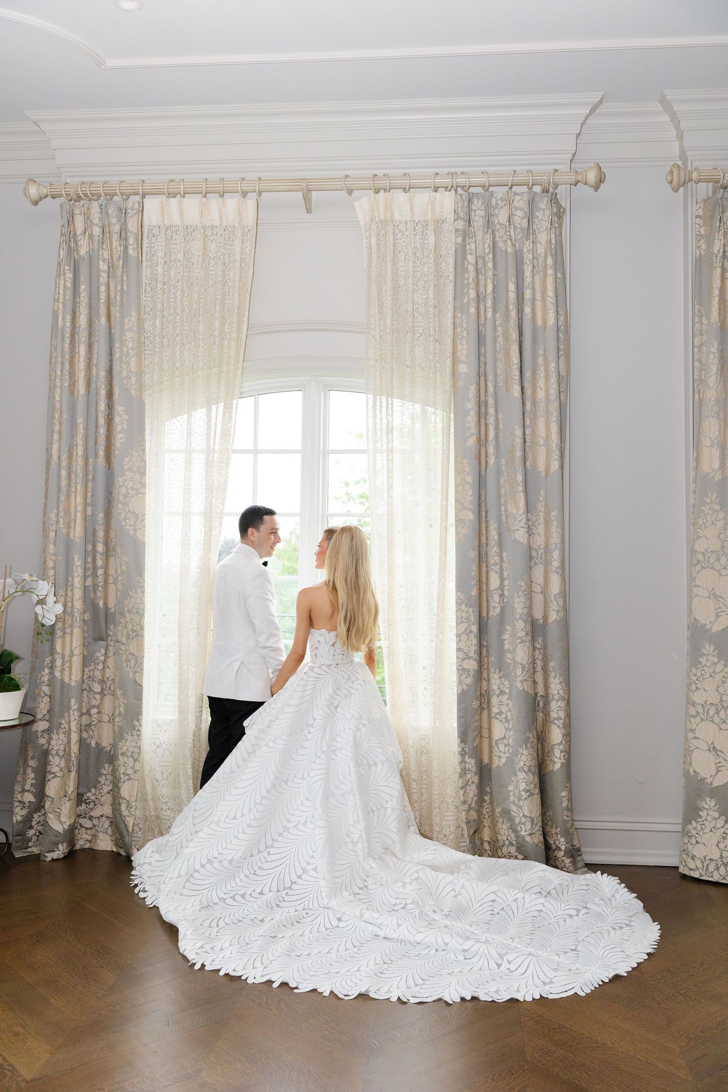 A flowing white wedding gown with intricate lace details and cathedral train displayed in a bright room with elegant curtains.