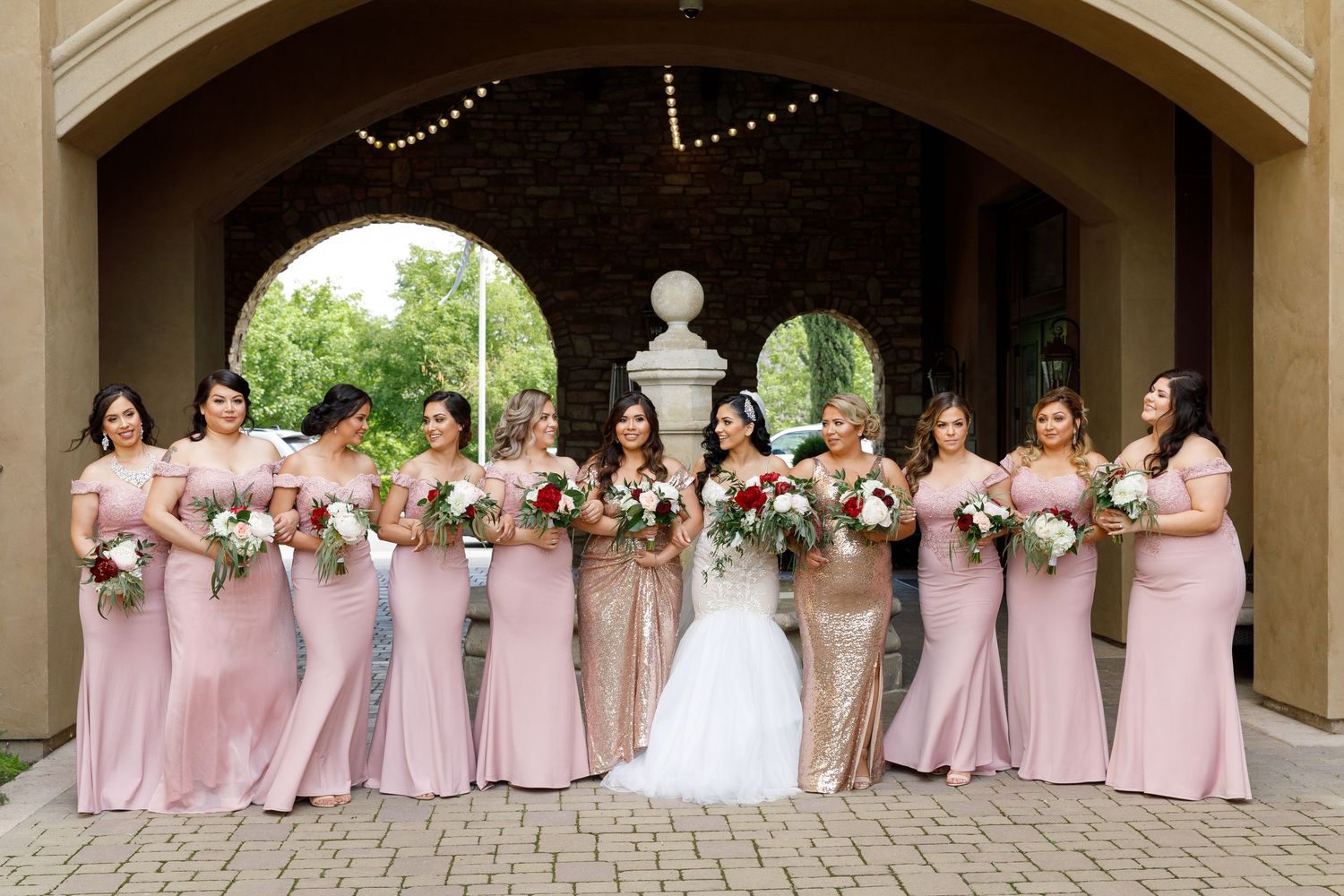 Wedding party wearing pink bridesmaid dresses poses together under an elegant covered archway.