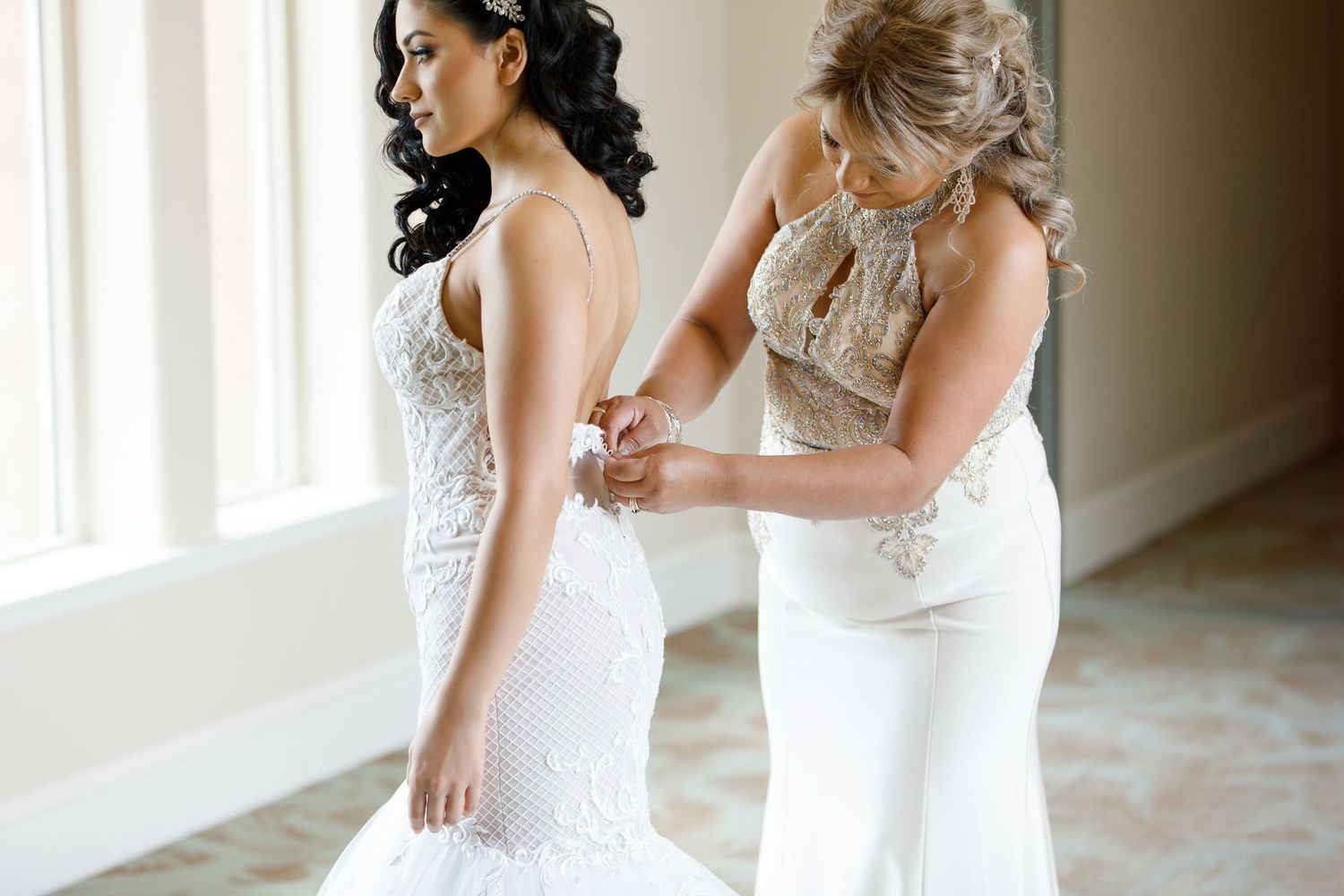 A bride in a lace wedding dress gets help with final adjustments in a sunlit room before the ceremony.