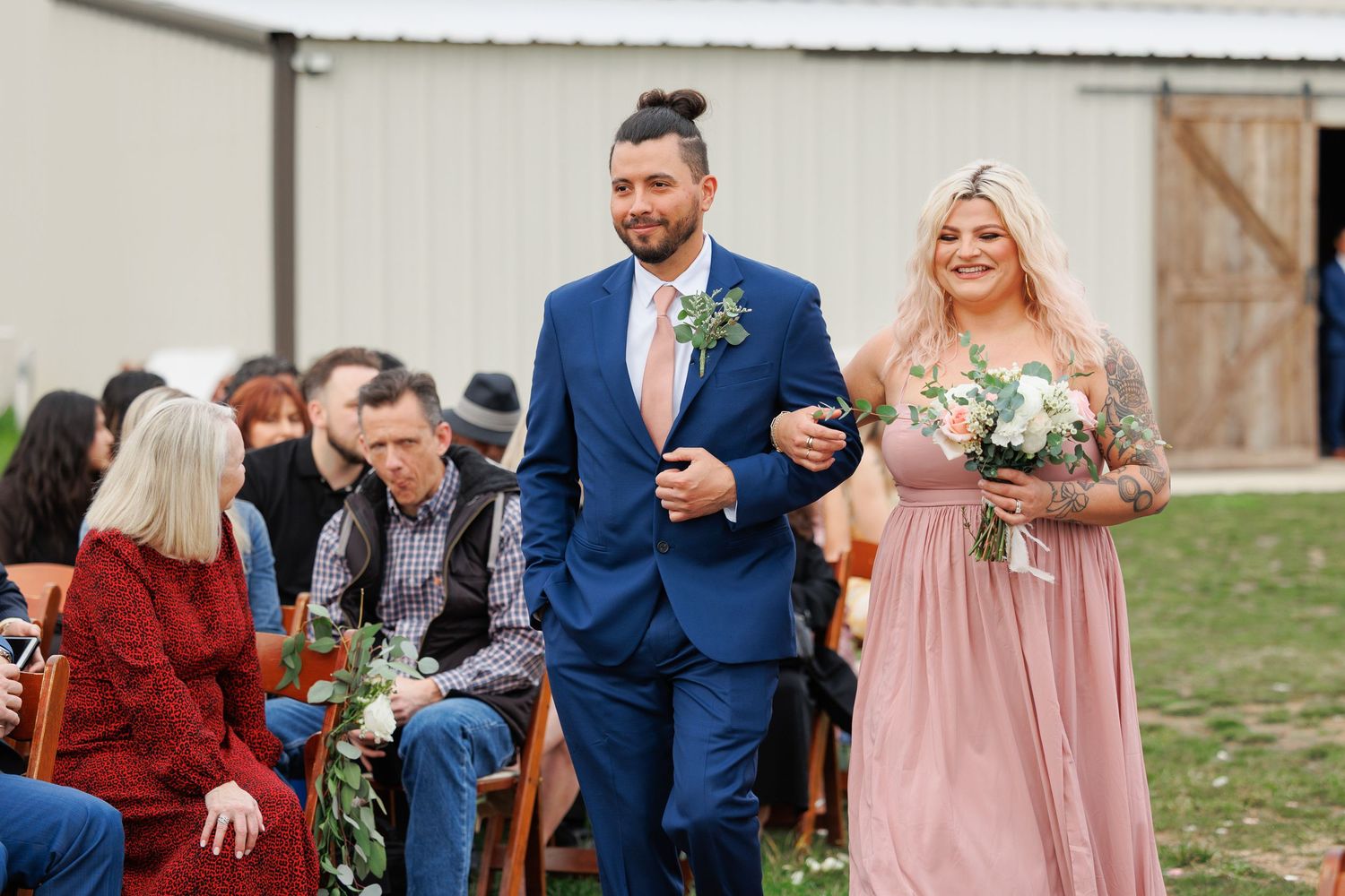 A joyful wedding processional with attendants in navy blue and blush pink walking down the outdoor aisle.