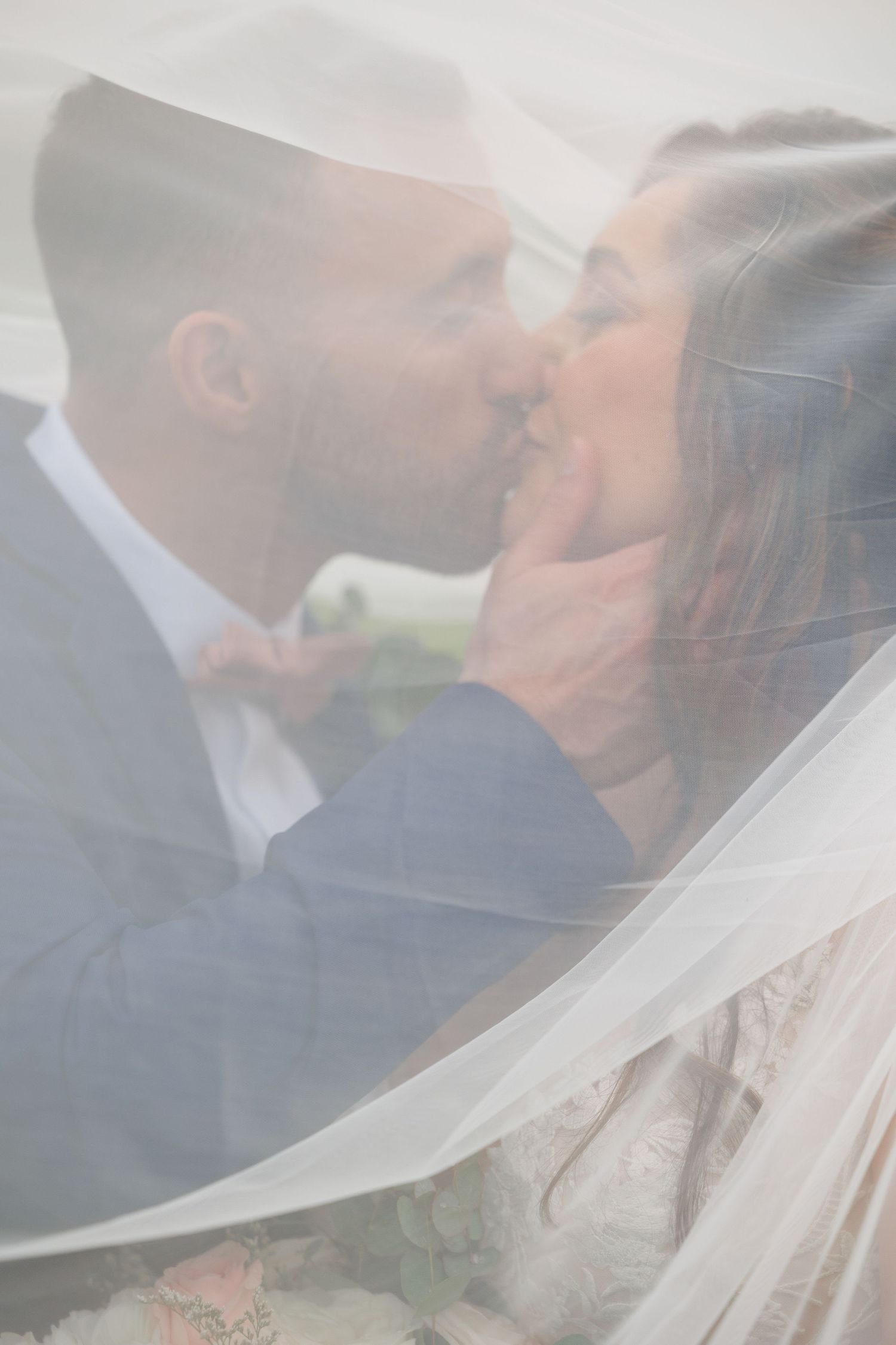 A romantic wedding photo series showing a couple kissing through a flowing white veil in an outdoor setting.