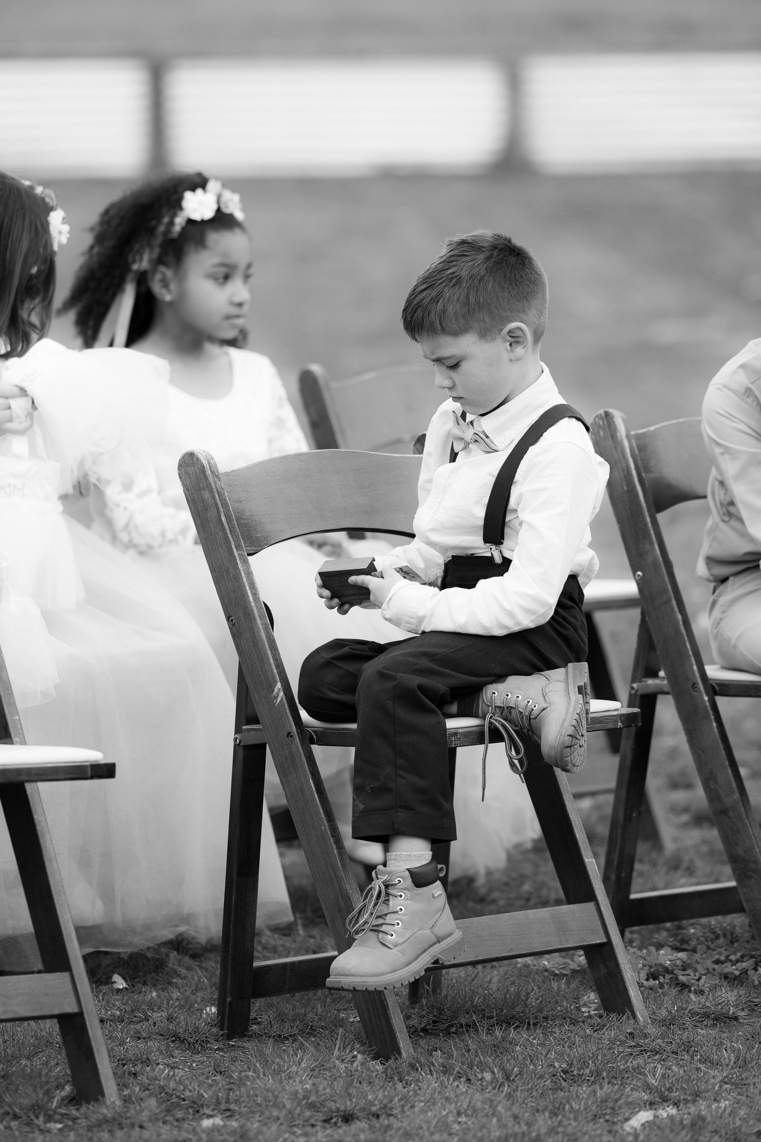 Black and white photo of young children in formal attire sitting on wooden chairs at an outdoor event.