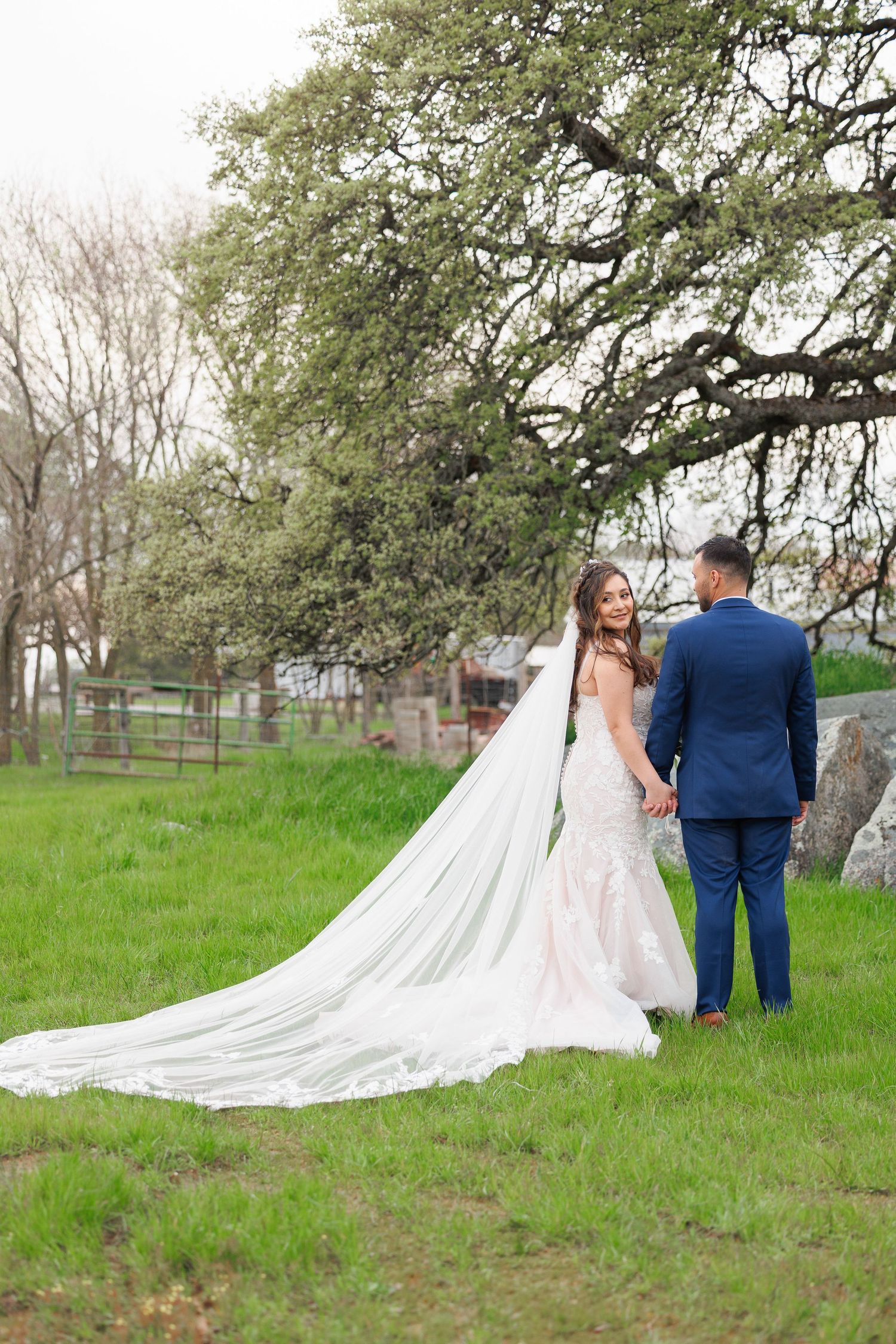 A bride and groom walk hand in hand through a grassy field with oak trees on a spring day.