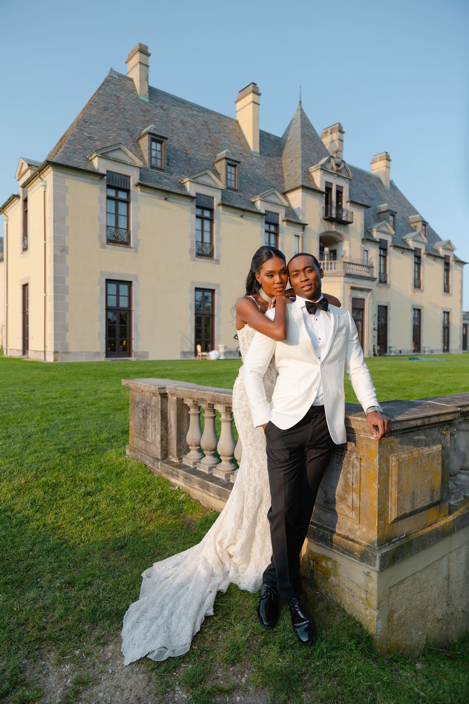 A wedding couple poses elegantly against a grand French chateau with stone balustrades and manicured lawn.
