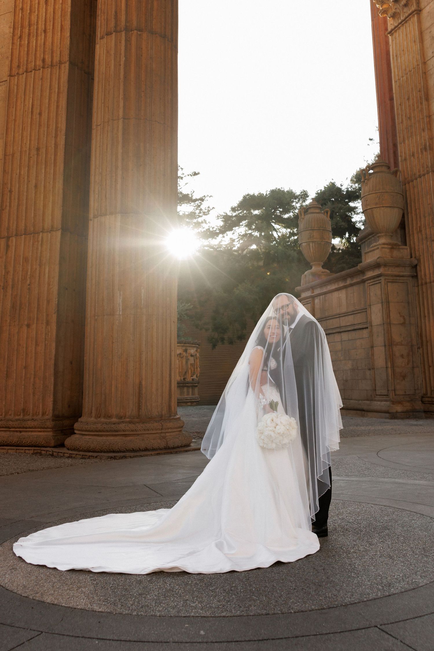 A bride in a flowing white gown and veil poses between classical columns at sunset.