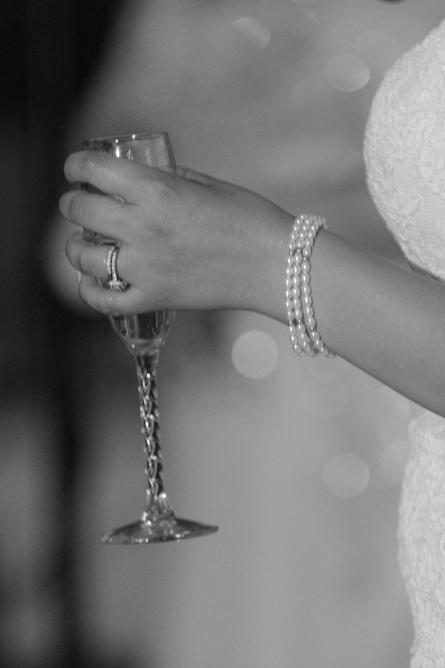 Black and white close-up of hands holding champagne glass with pearl bracelet detail.