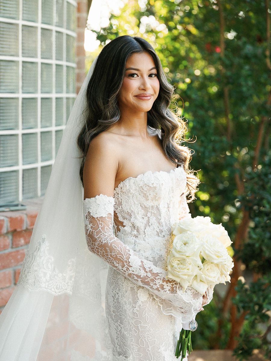 A bride in an off-shoulder lace wedding gown with long sleeves holds a white bouquet against a garden backdrop.