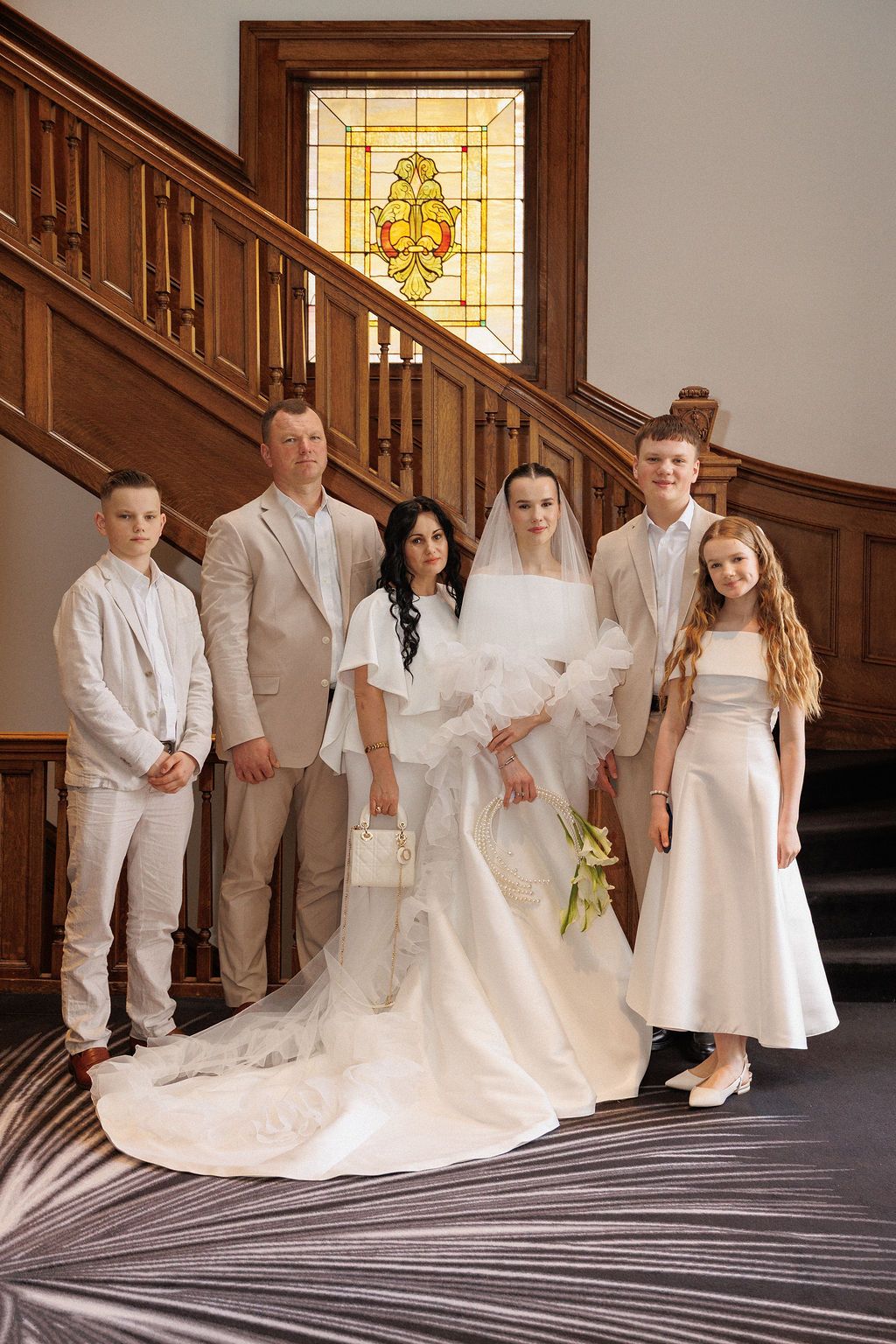 A wedding party in white and beige attire poses on an elegant wooden staircase with stained glass window backdrop.