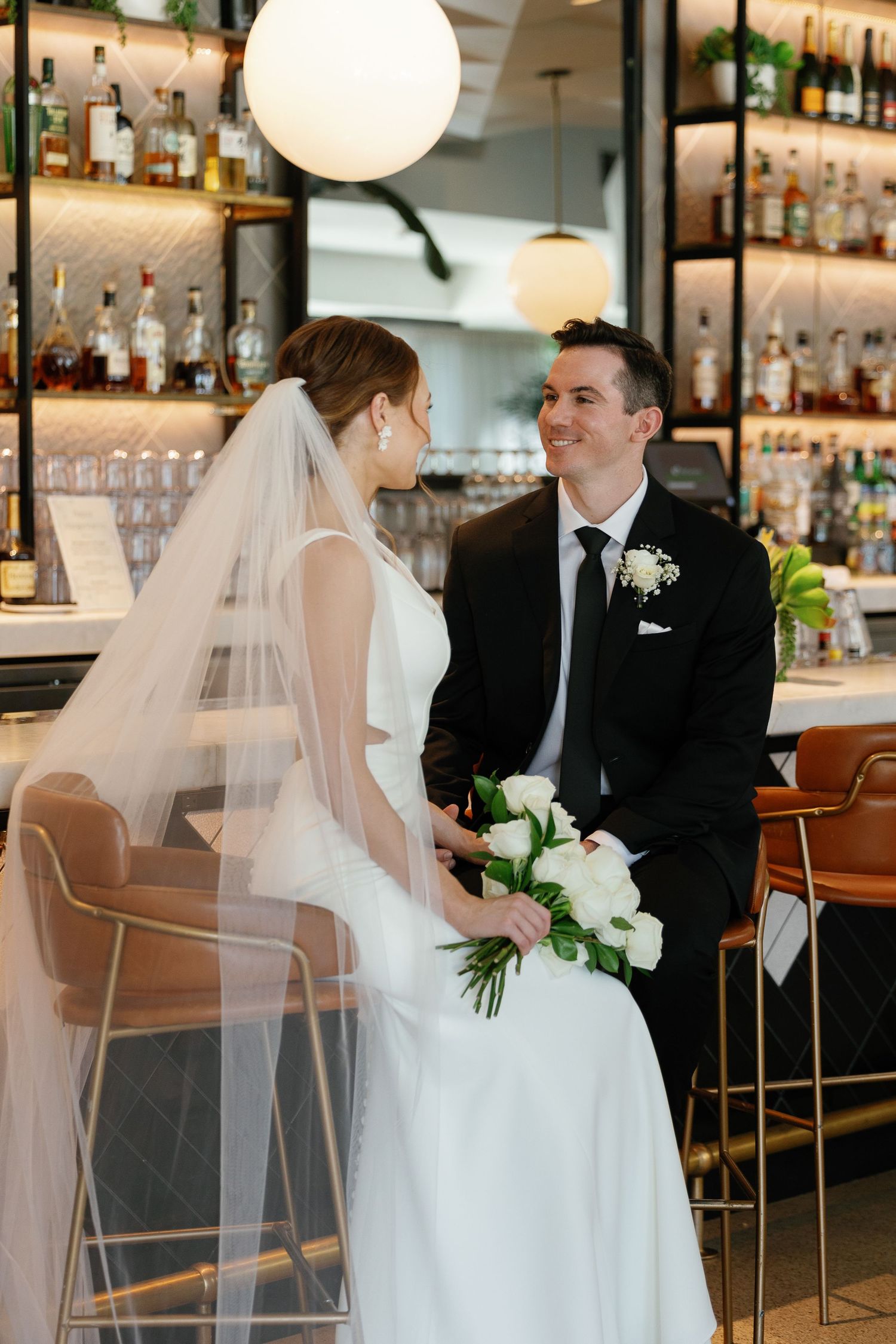 Newlywed couple shares intimate moments at an elegant restaurant bar with white decor and ambient lighting.