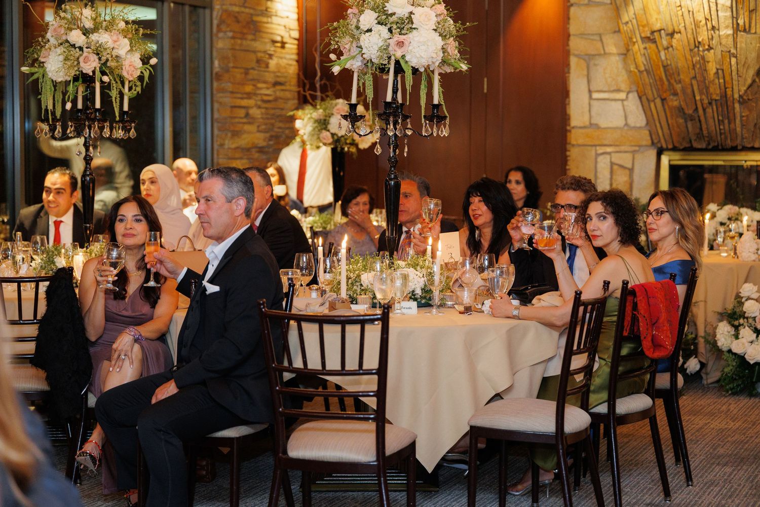 Wedding guests seated at elegant tables with chandeliers and floral centerpieces enjoy a reception celebration at an indoor venue.