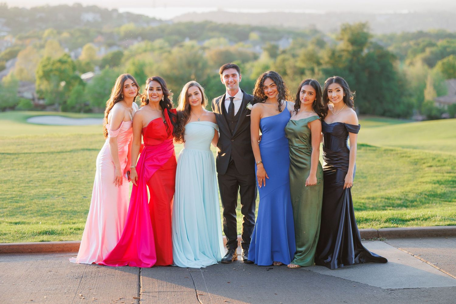 A group in formal evening attire poses together on a paved area overlooking a scenic green landscape at sunset.