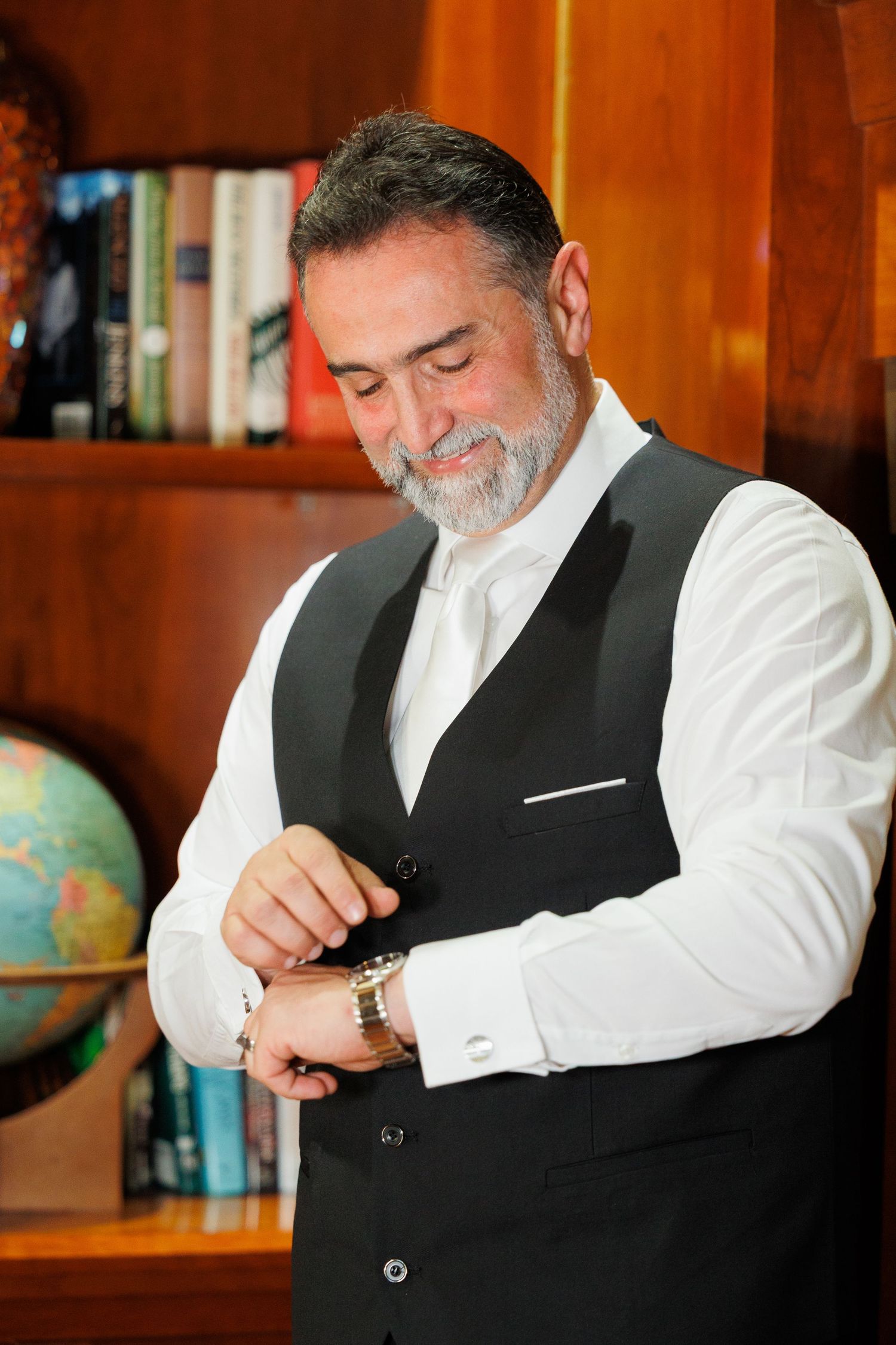 A person in a black vest and white shirt adjusts their watch in front of wooden bookshelves with a globe visible.