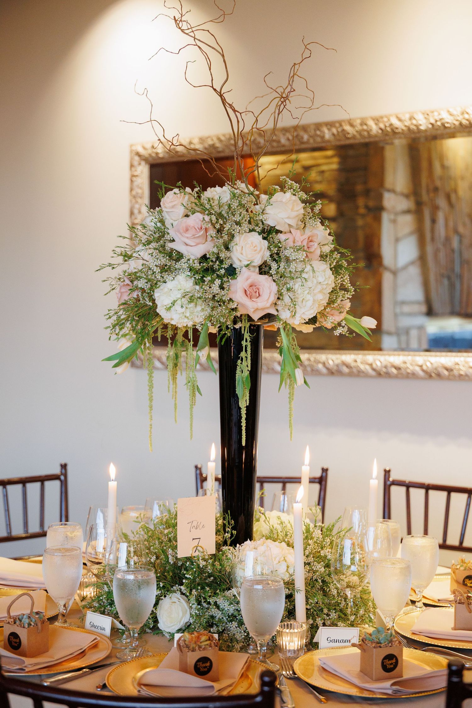 Elegant wedding reception table setting with tall black vases filled with pink roses and greenery, surrounded by glowing candles.