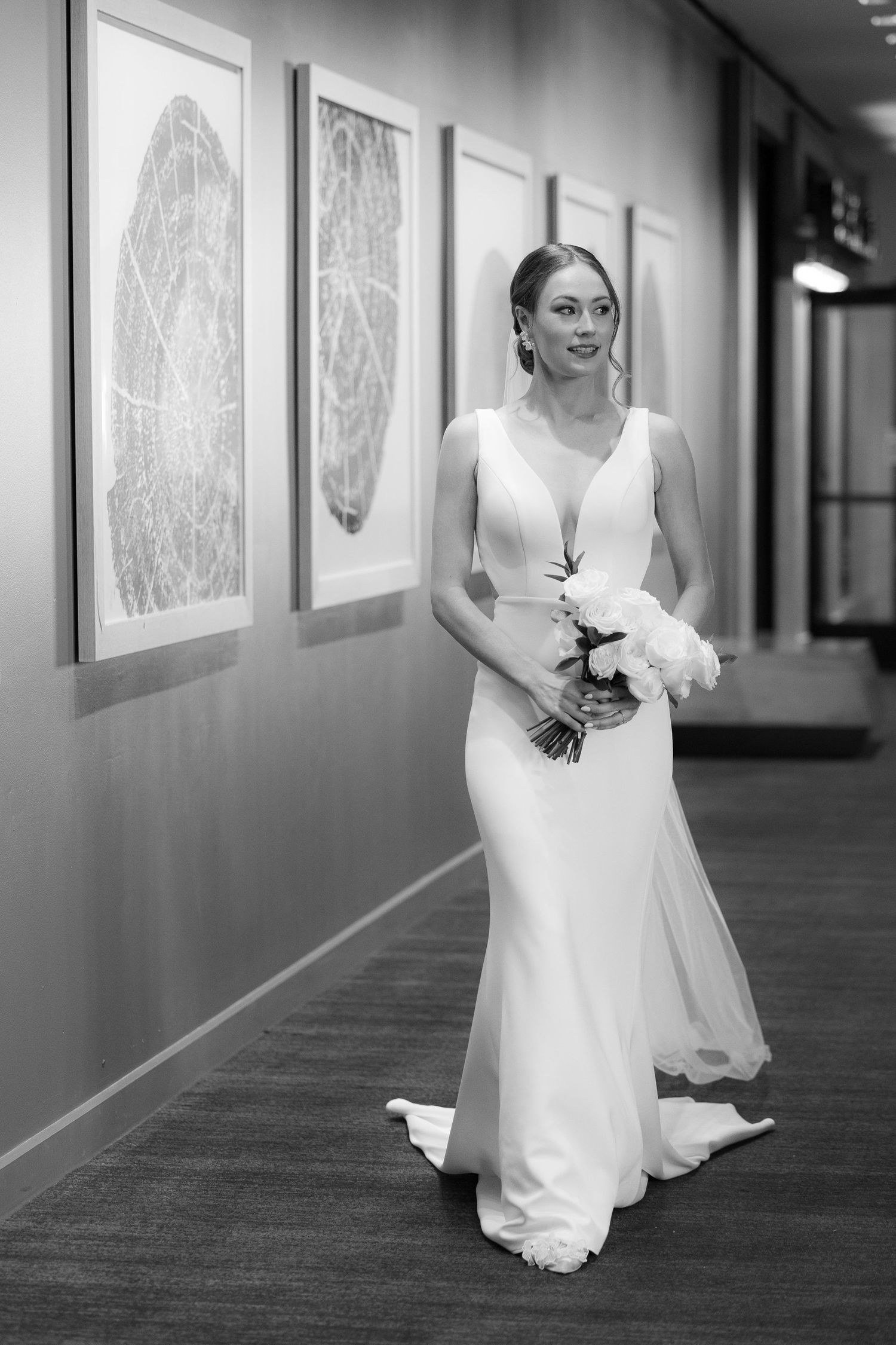 A bride in a white wedding gown walks down a hotel hallway with framed artwork on the walls.