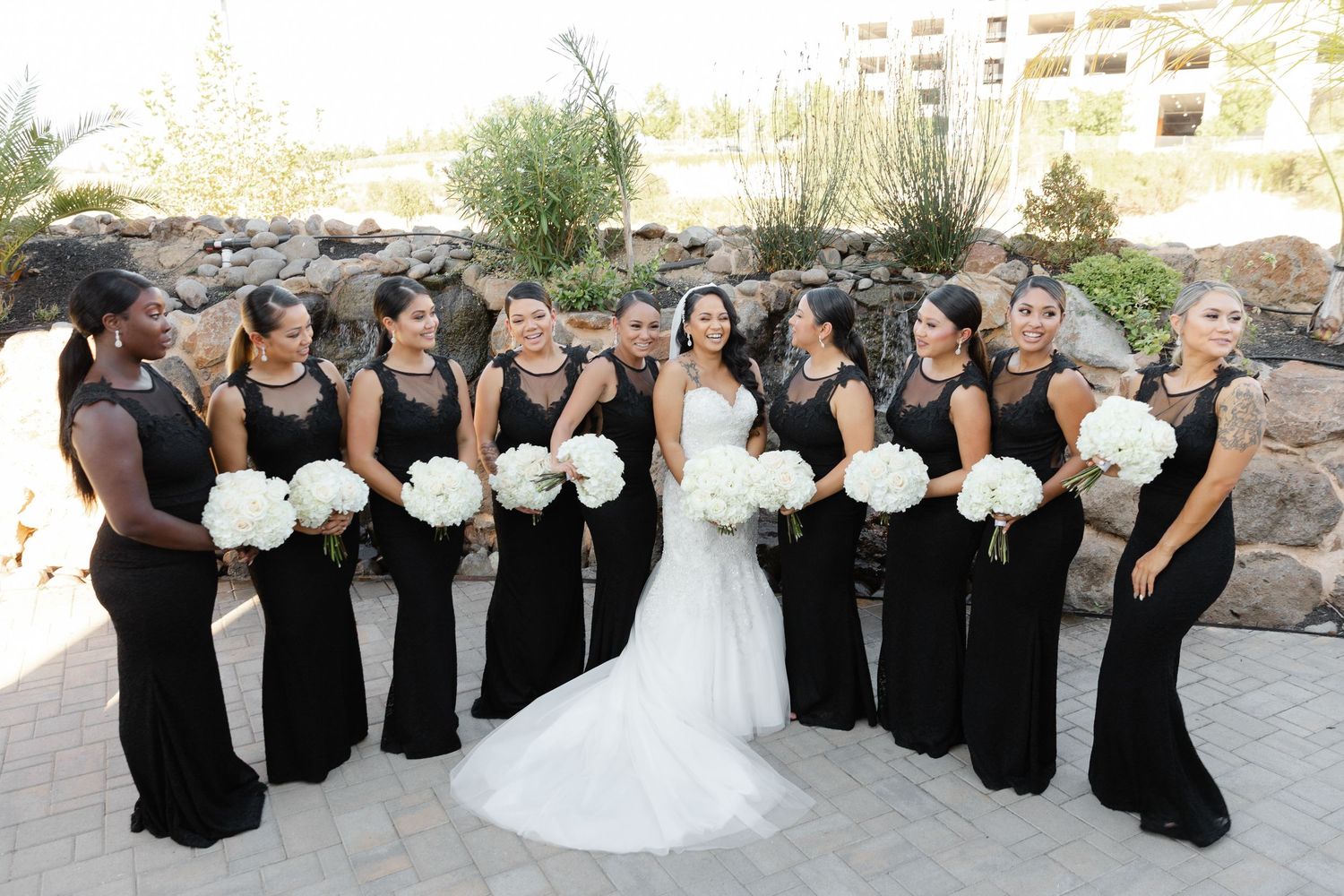 Wedding party in black dresses and white gowns pose together outdoors on stone steps for formal group photos.