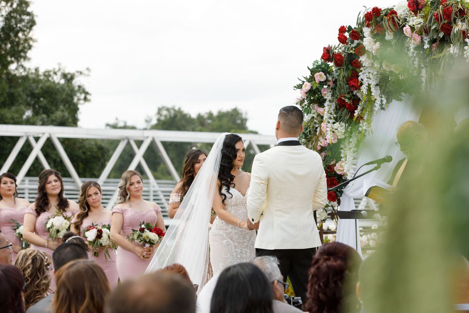 Wedding ceremony setup with white chairs and floral arrangements under trees at an outdoor venue.