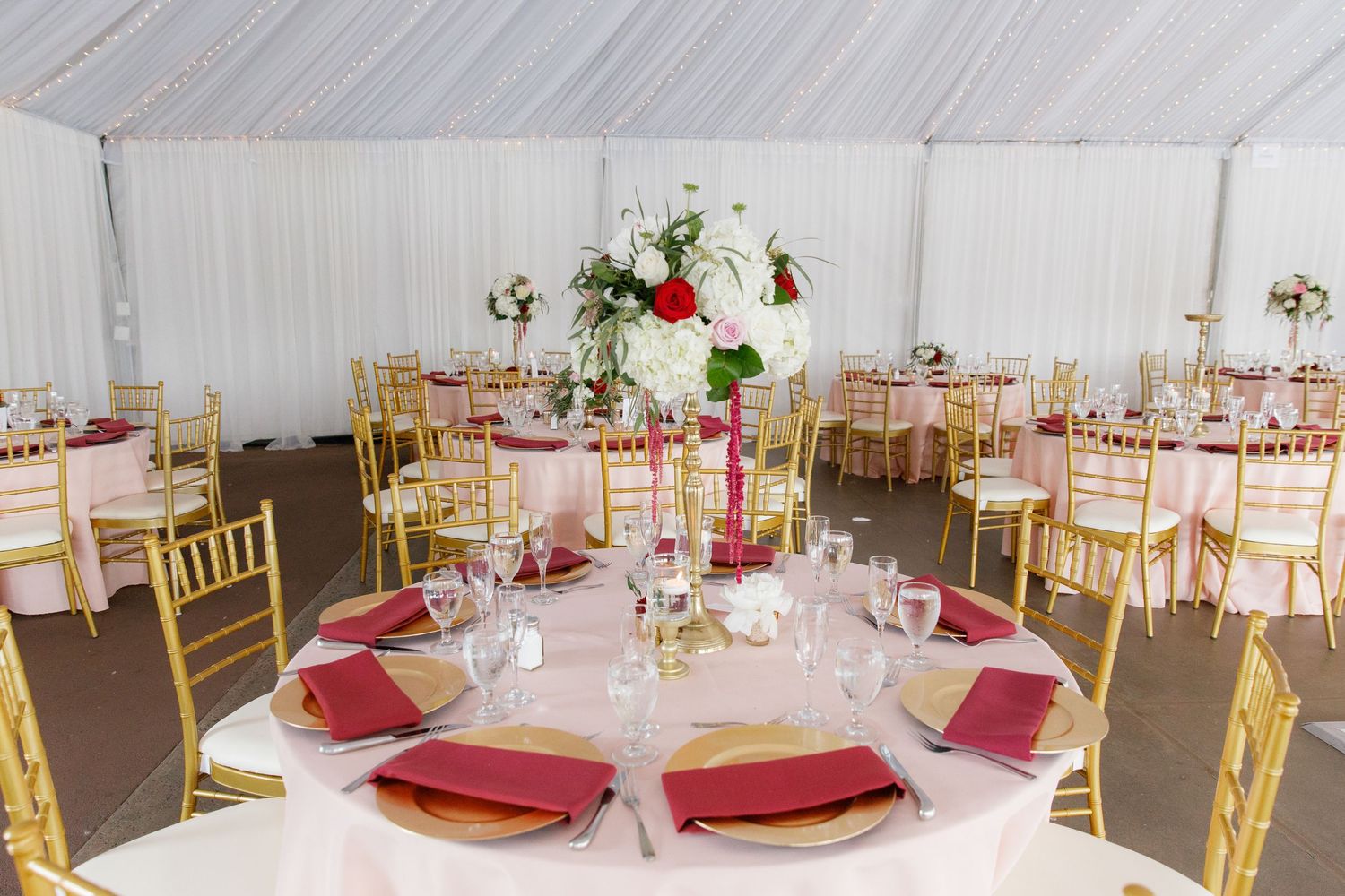 Elegant wedding reception tables with gold chairs, burgundy napkins and white floral centerpieces in a white tent setting.