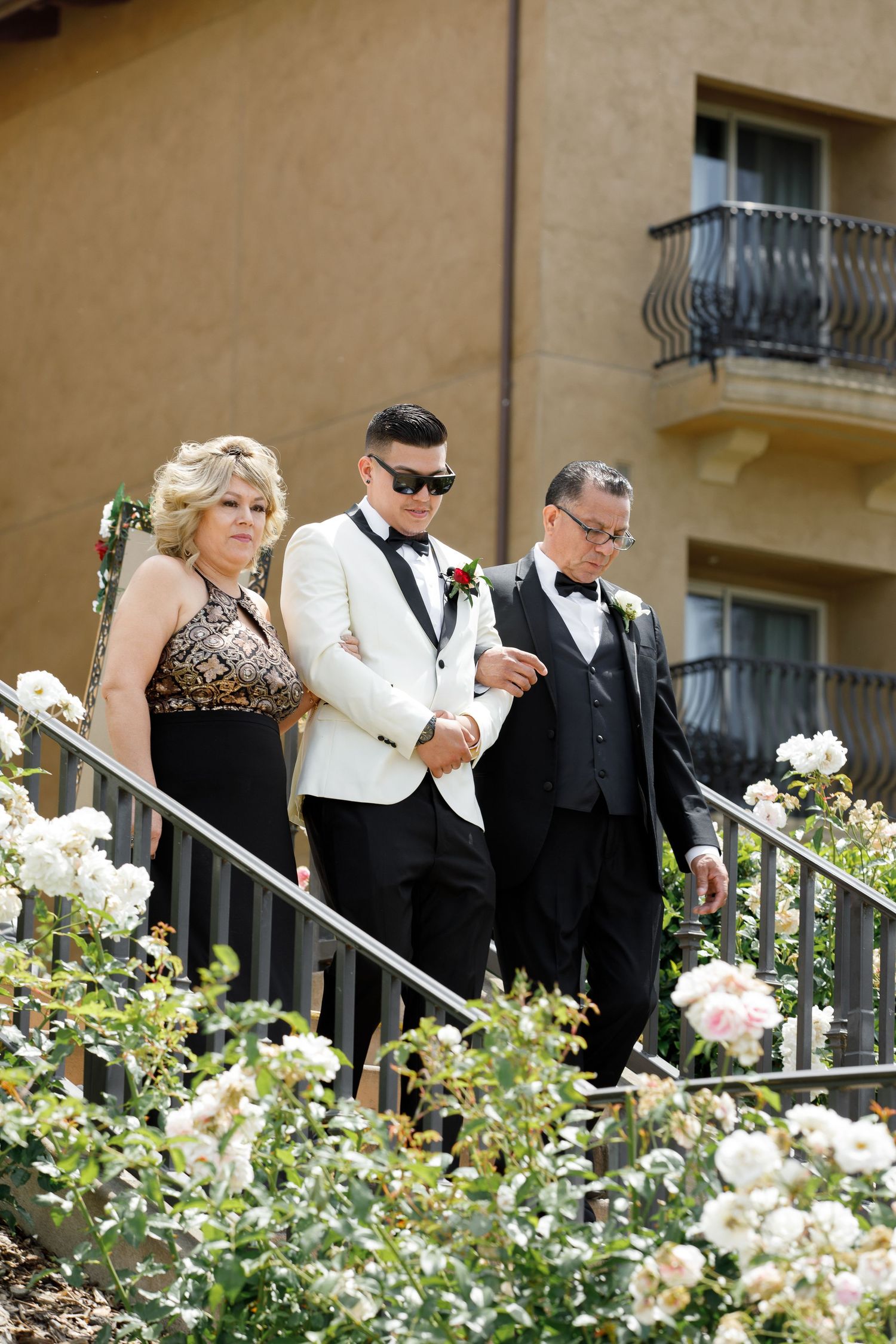 Three people in formal attire descend stairs bordered by blooming white roses at an elegant venue.