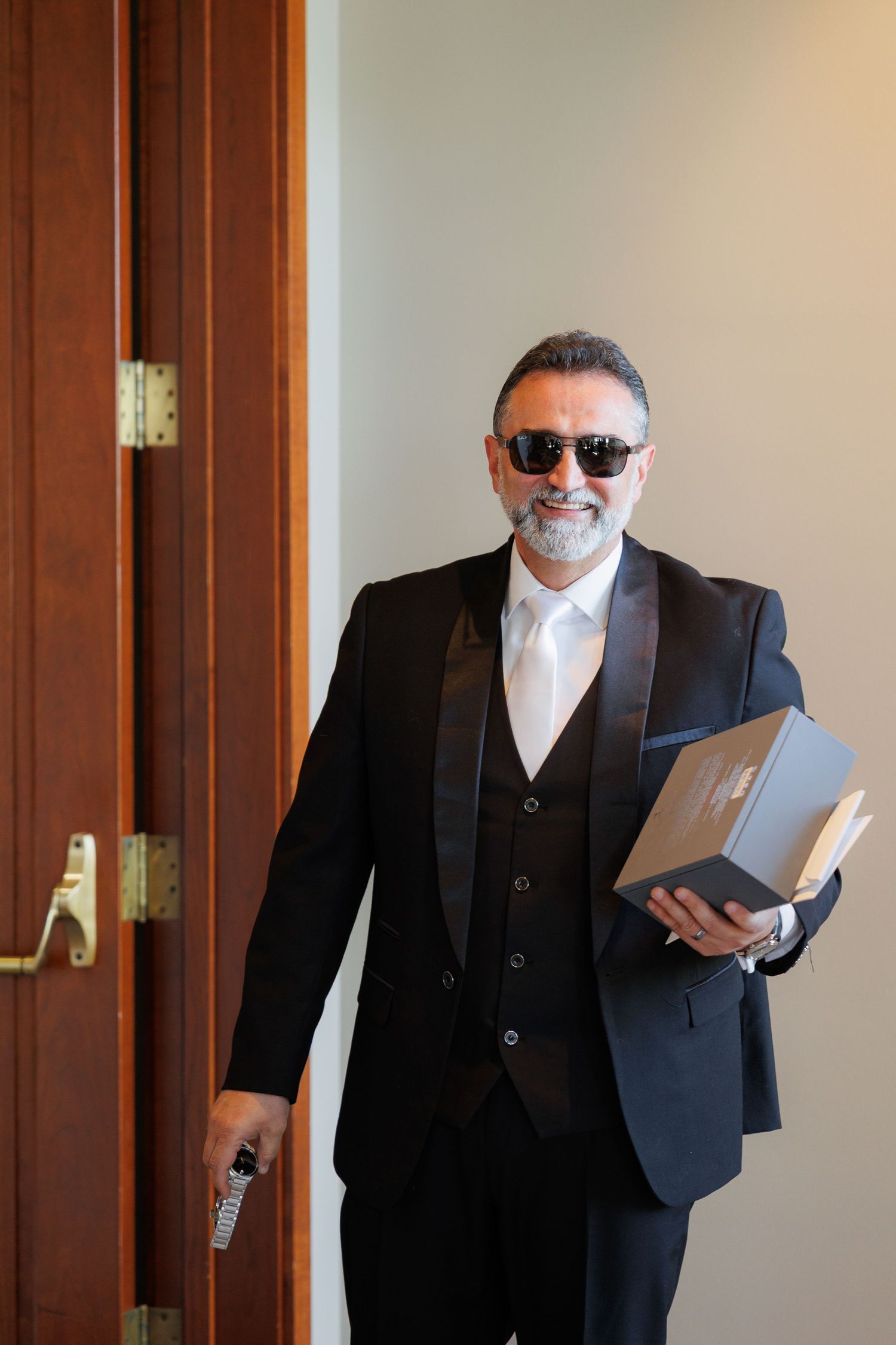 A person in a black suit and sunglasses walks down a hallway while carrying a laptop or tablet device.