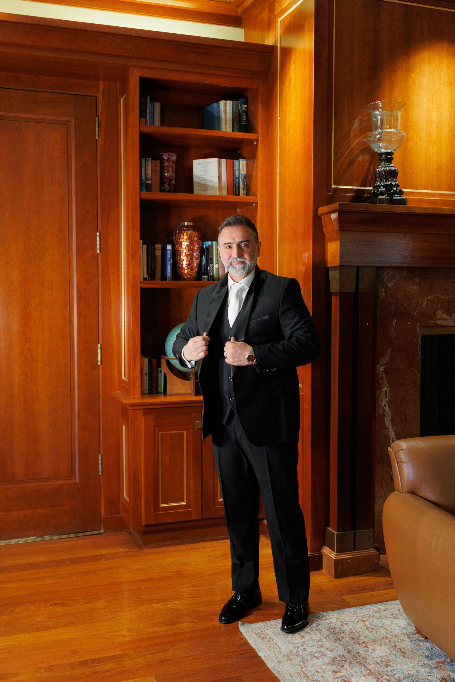 Person in black suit strikes playful poses in elegant wood-paneled room with hardwood floors and built-in cabinets.