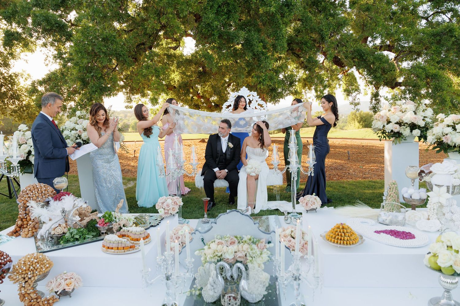 An elegant outdoor wedding reception features long tables decorated with floral centerpieces under a canopy of trees at sunset.