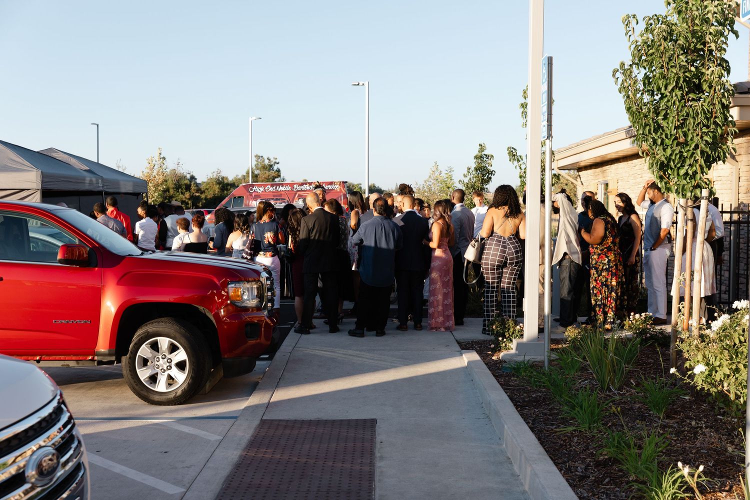 A panoramic view of a parking lot with cars and people gathered outside a building during sunset.
