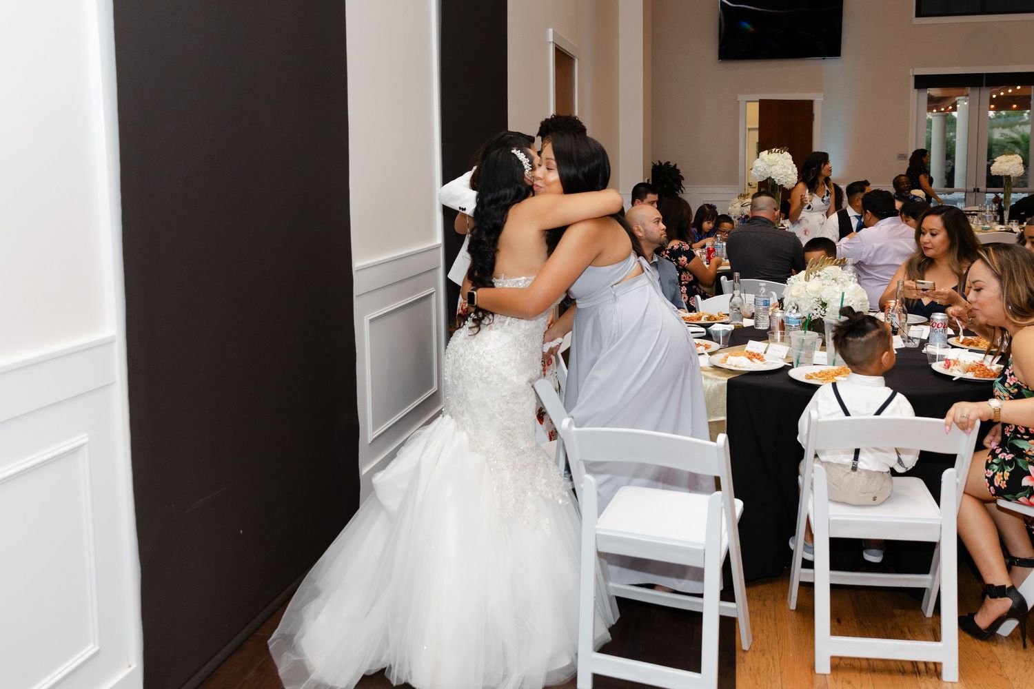 Bride in white wedding dress sits at a table during a reception while guests gather in the background.