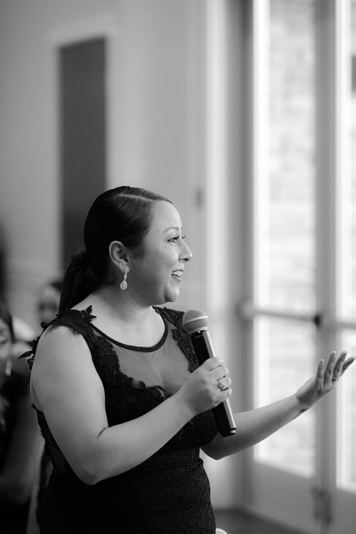 A speaker in a black dress gestures while giving a presentation with a microphone in a naturally lit room.