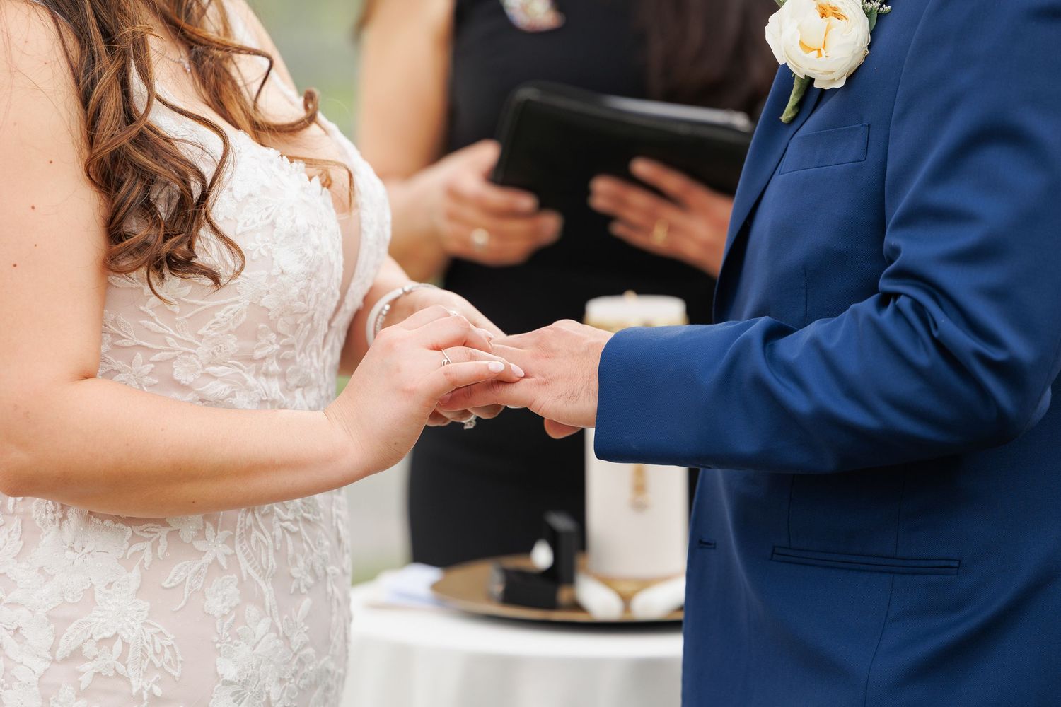 A close-up of hands exchanging wedding rings during a ceremony, with the bride wearing a white lace dress.