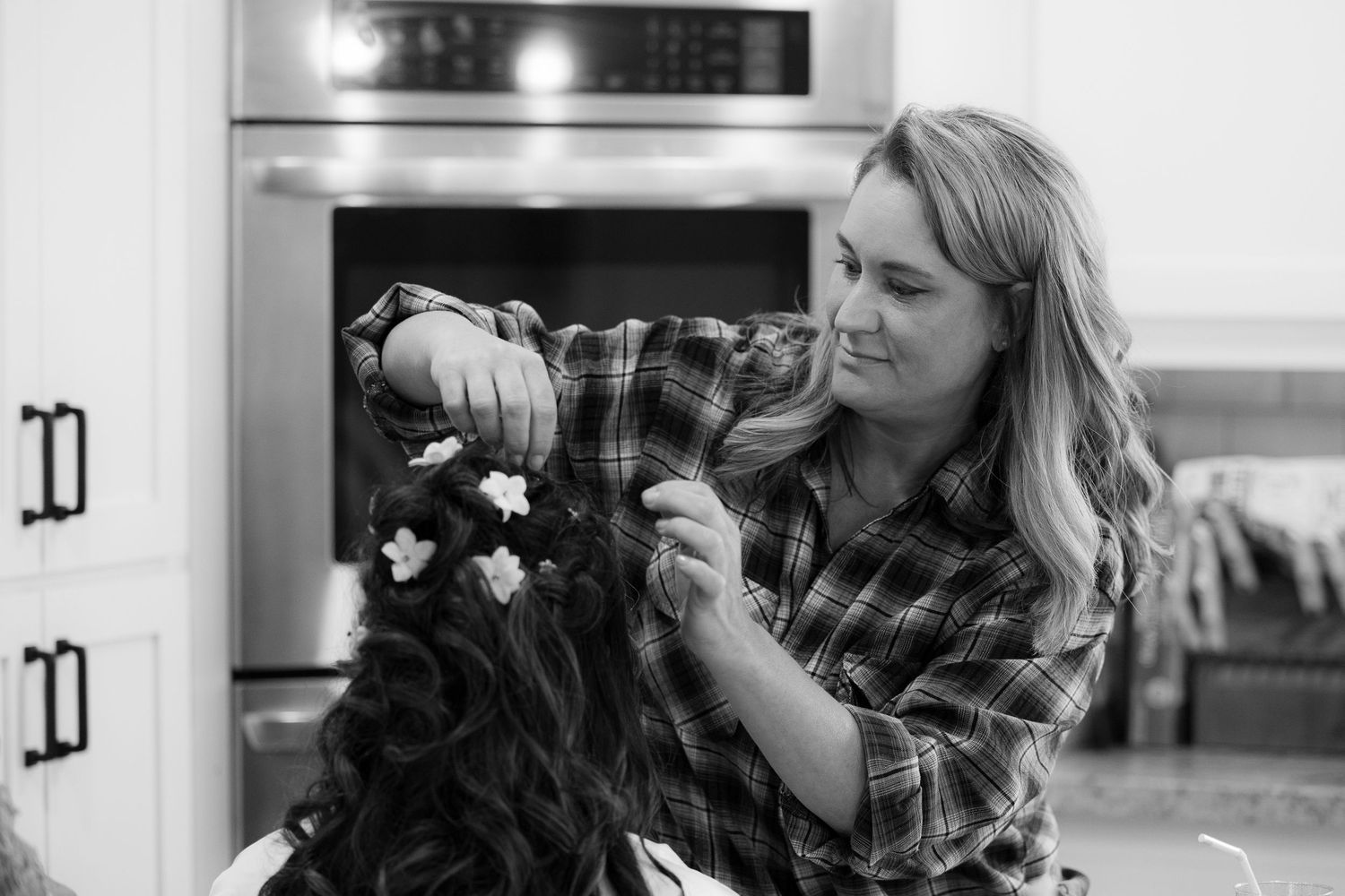 A hairstylist wearing a plaid shirt styles curly hair in a salon setting, captured in black and white photography.