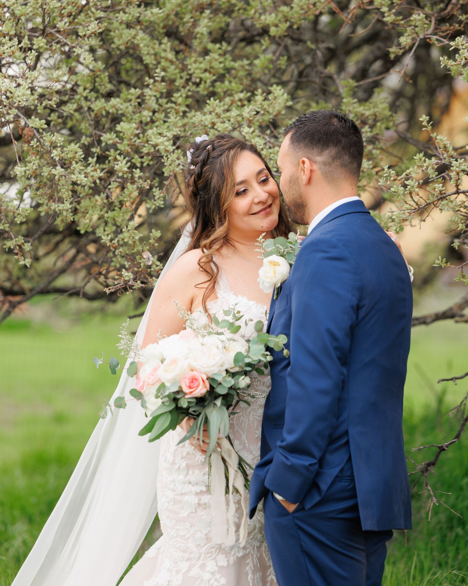 A romantic wedding couple poses together under blooming spring tree branches in an outdoor garden setting.