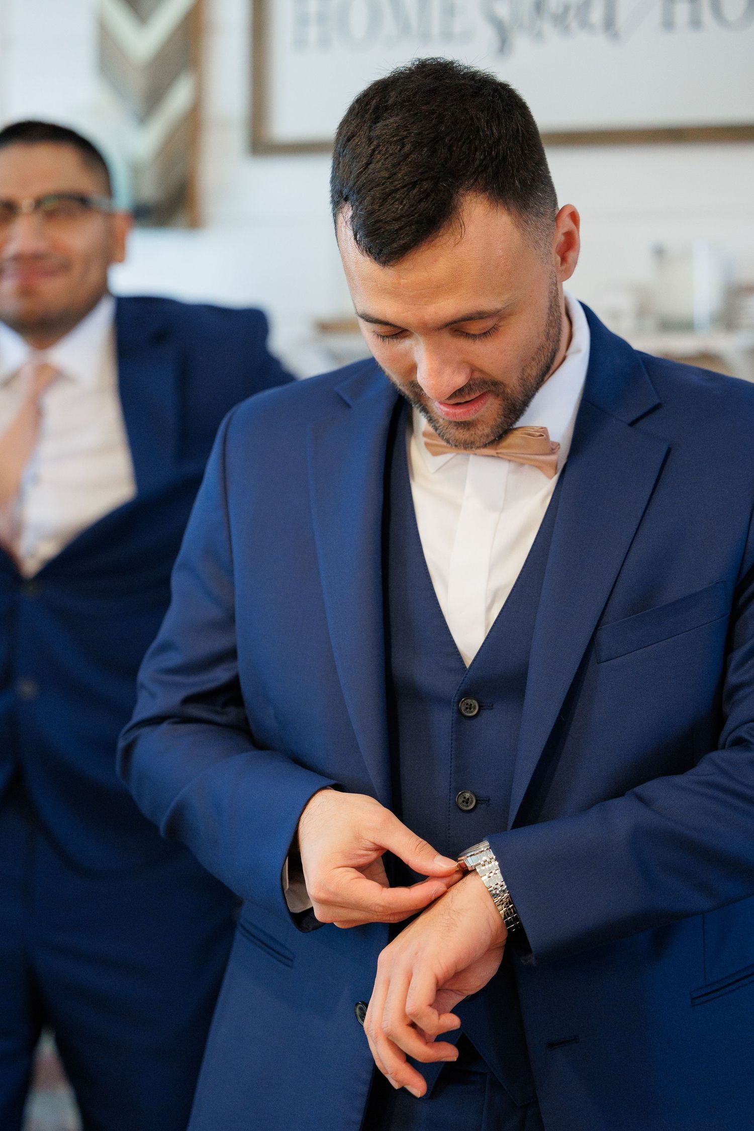 A groom in a navy three-piece suit checks his watch while getting ready for his wedding ceremony.
