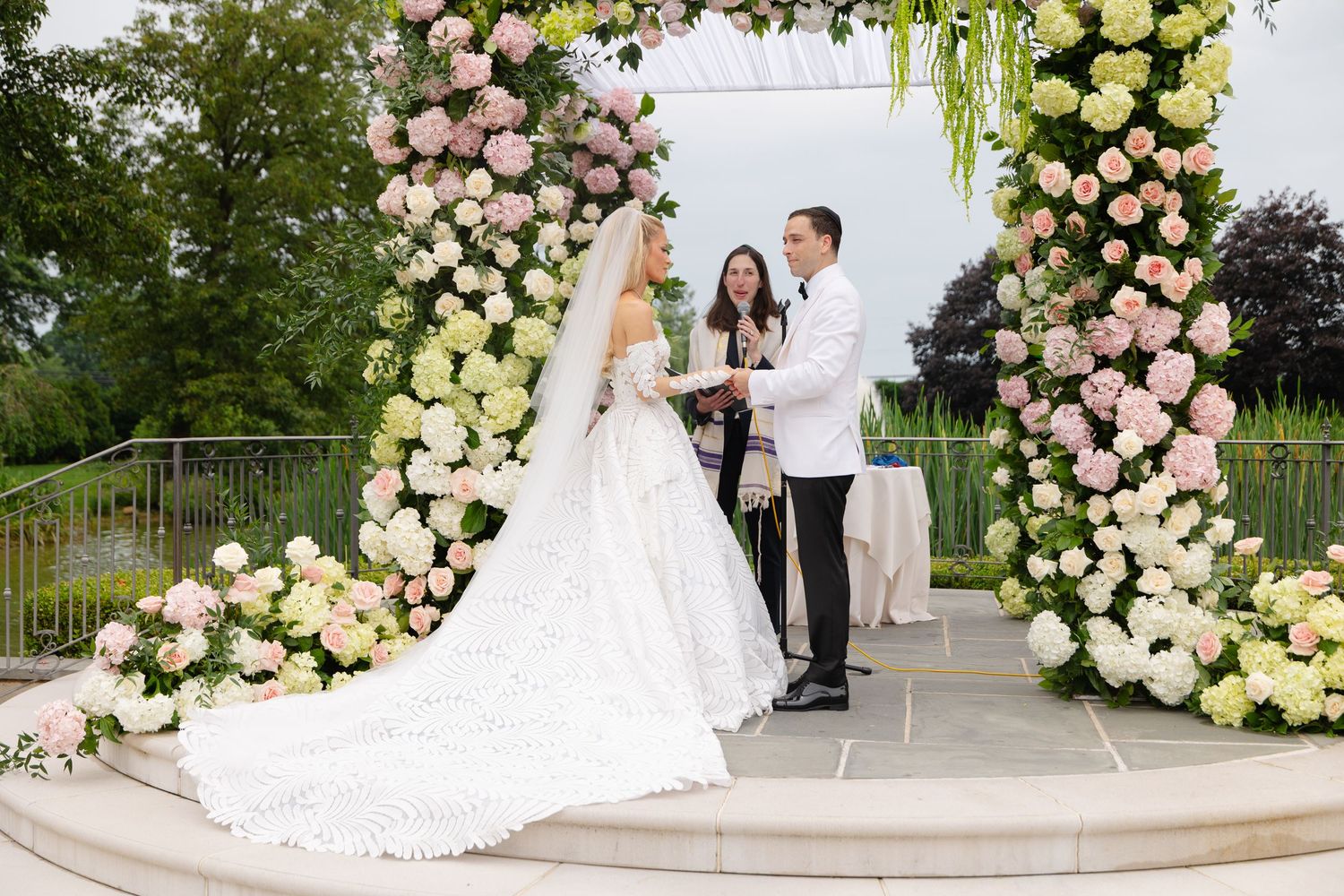 A wedding ceremony takes place under an elegant floral arch with pink and white roses on a stone patio.