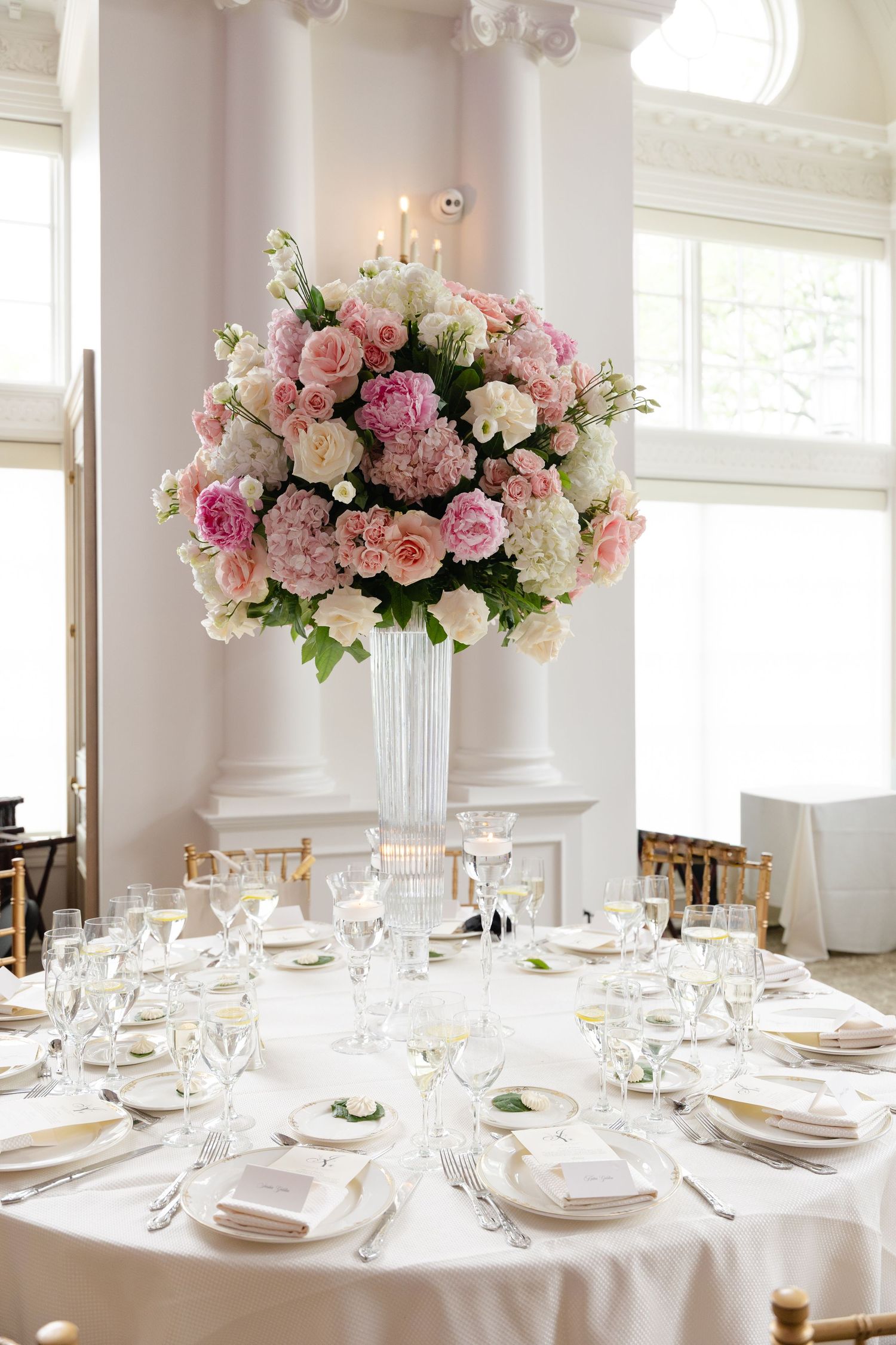 Elegant wedding reception tables set with white linens, china, and tall pink rose centerpieces in white vases.