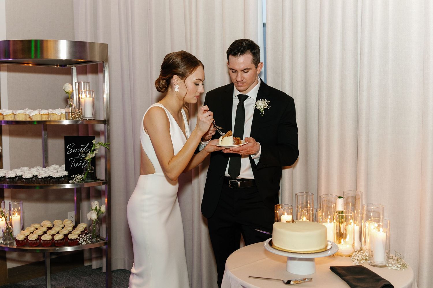 A couple shares cake at their wedding reception with candlelit dessert table and shelving display in background.