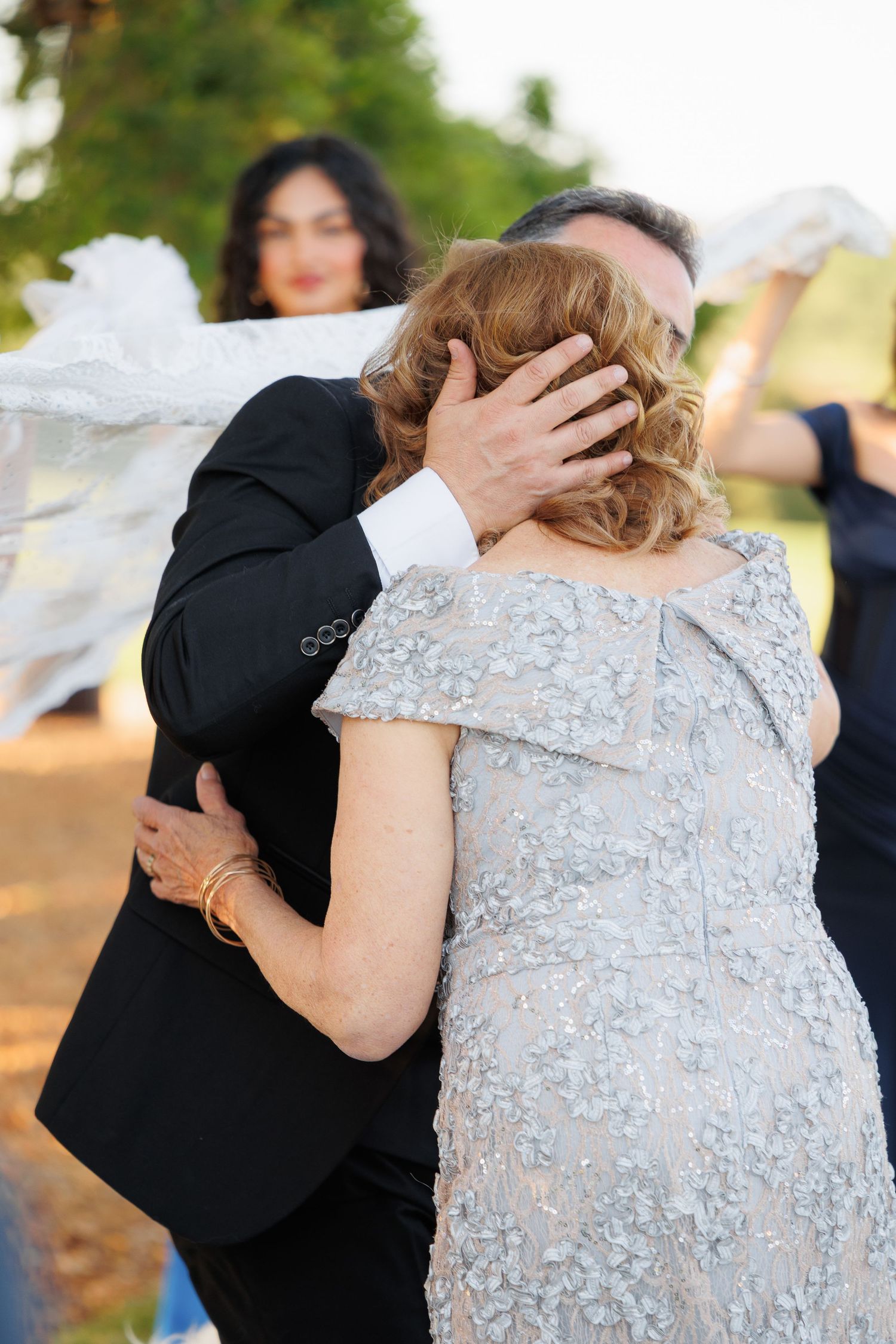 A couple embraces during a wedding ceremony, with the bride wearing a beaded silver gown and groom in black suit.