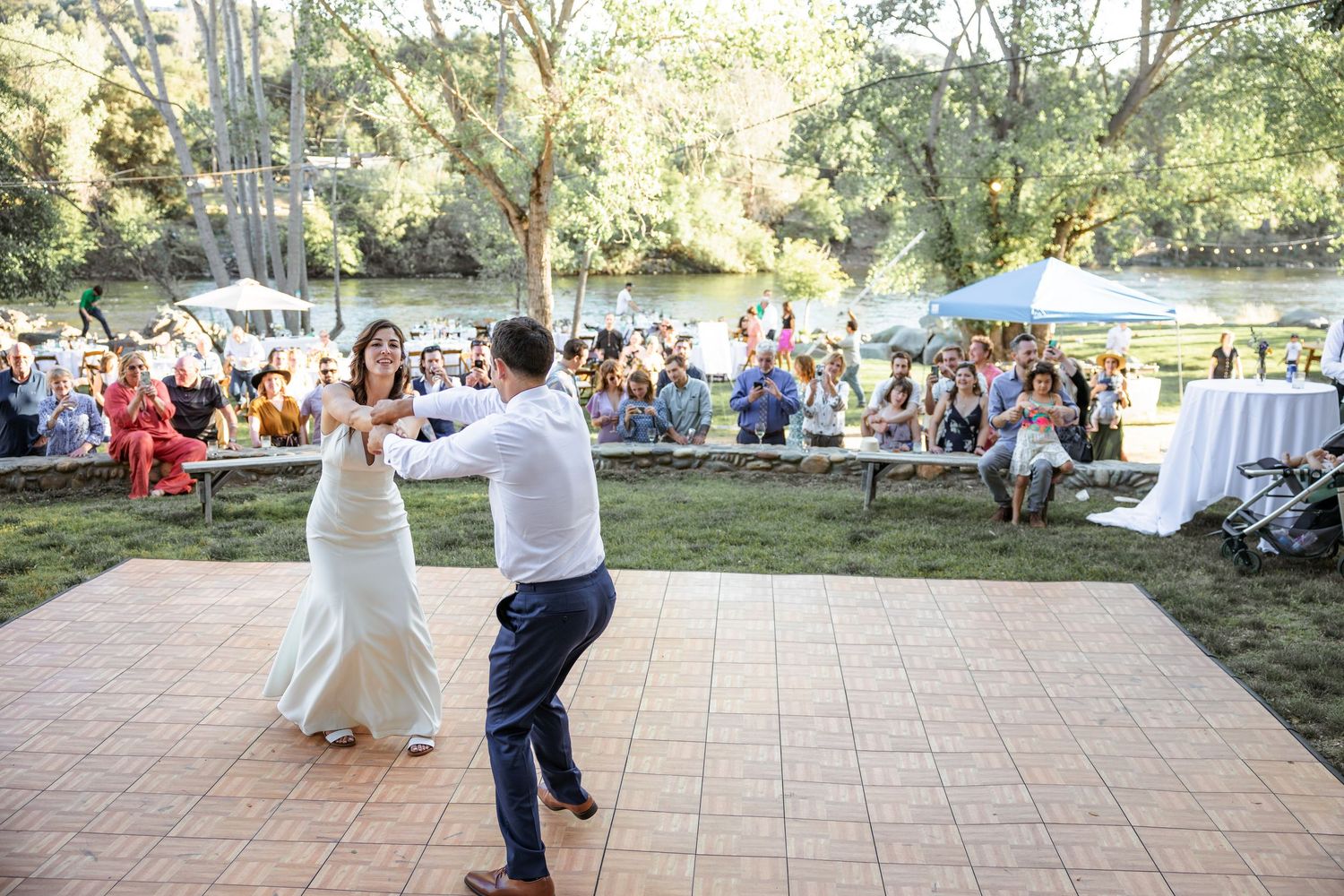 A couple shares their first dance on an outdoor patio while wedding guests watch from the lawn during a summer celebration.