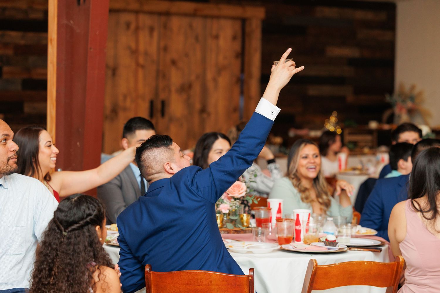 Wedding guests raise their hands in celebration at a rustic wooden barn reception venue during dinner.
